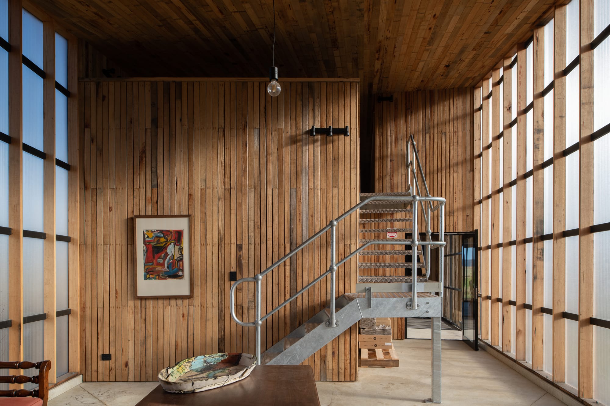 Tasmanian Timber Series: Westella Vineyard.Spacious interior of Westella Vineyard featuring a high ceiling with exposed beams, Tasmanian Oak walls, and a staircase leading to a loft area, complemented by natural light and a cozy sitting area.