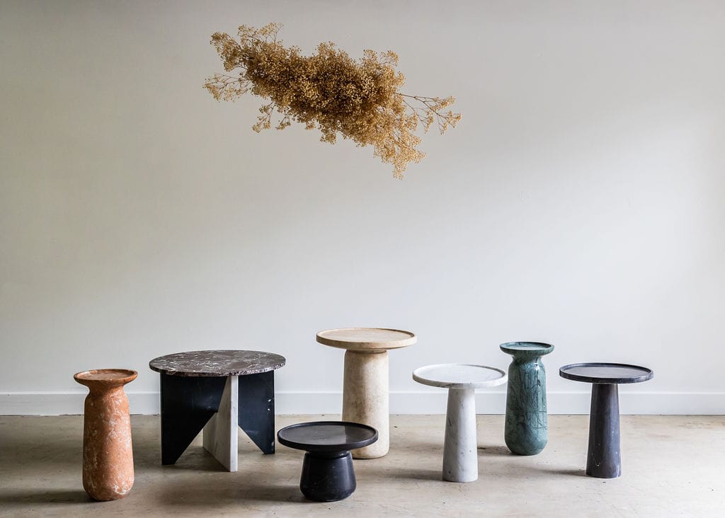 Noma Co Studio. Copyright of Noma Co Studio. Arrangement of different round, marble end tables in a range of beige, orange, white and blue tones. Concrete floor, white walls.