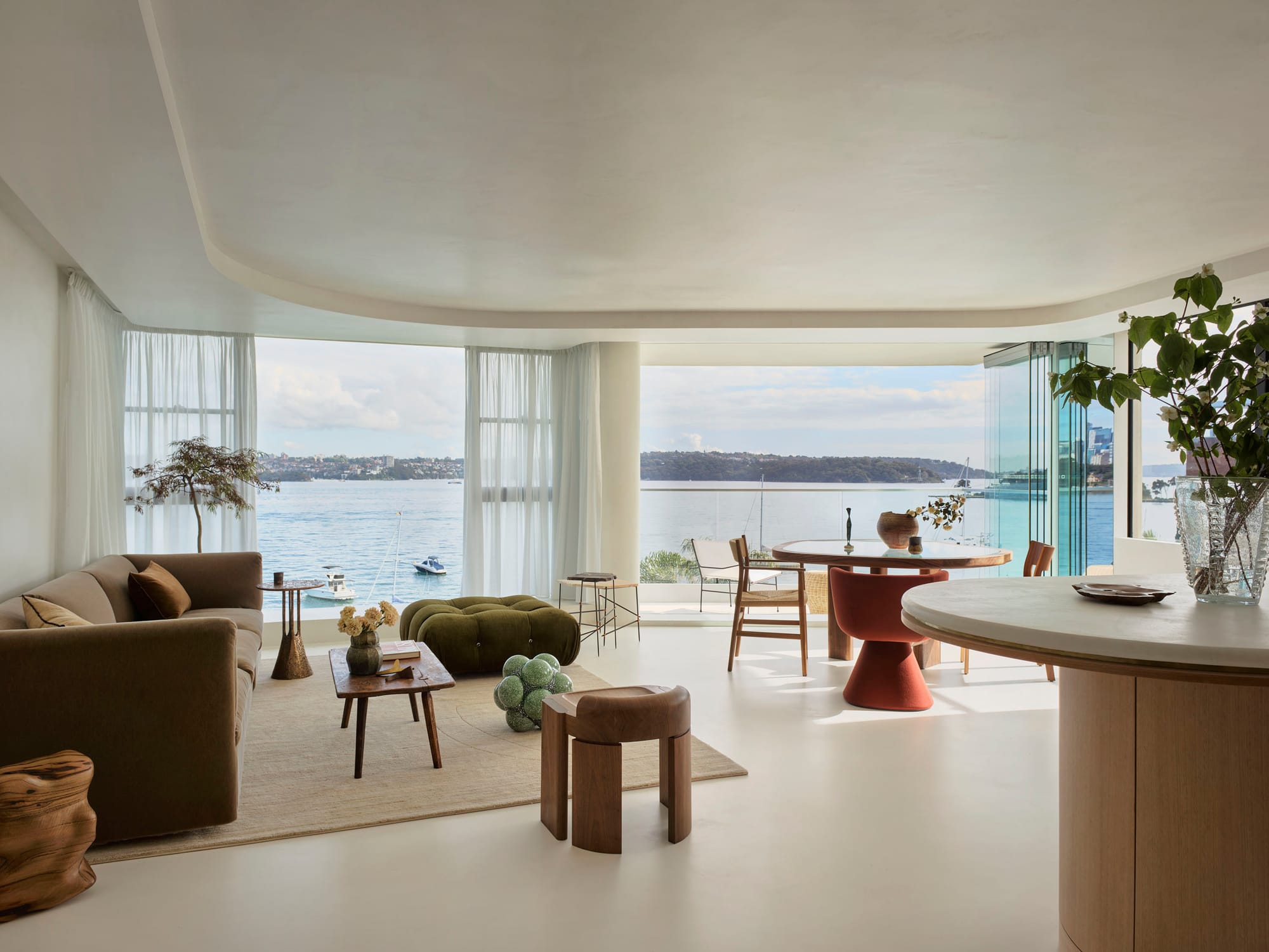 Tasmanian Timber Series: Darling Point Apartment. Photography by Ansen Smart. Wide angle shot of open plan, modern living space in residential apartment. Floor to ceiling windows in background of image open up to show harbour views of water and hills. Floors and walls finished in neutral white plaster. Organically curved recess integrated into ceiling. Floor to ceiling white sheer curtains over windows. Curved Tasmanian Oak kitchen counter in right hand foreground of image. Eclectic furniture scattered around space.