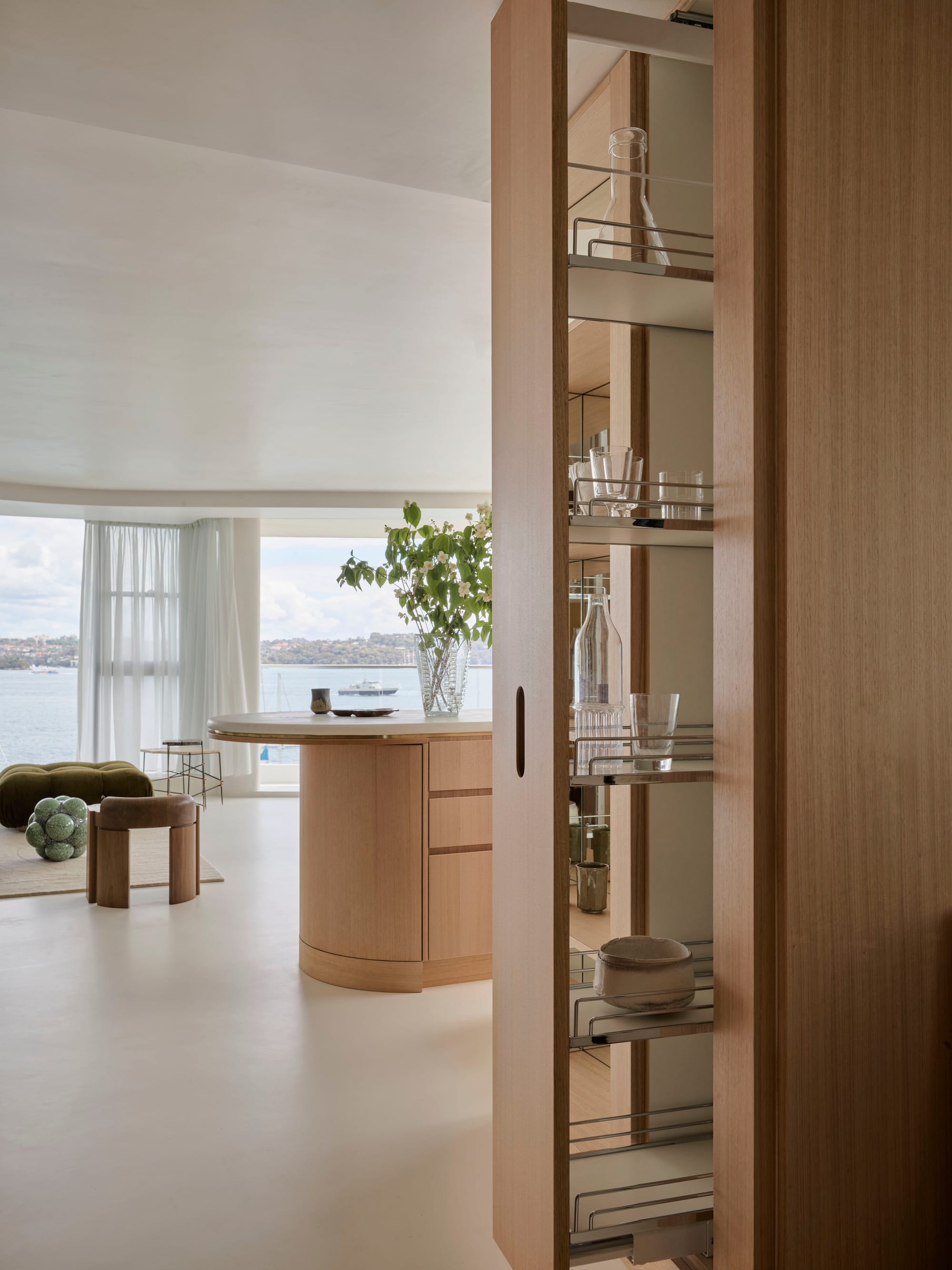 Tasmanian Timber Series: Darling Point Apartment. Photography by Ansen Smart. Detail image of integrated kitchen cabinetry in residential apartment space. Tasmanian Oak joinery for storage and organically curved island bench top, finished with white countertop. White flooring, walls and floors. Floor to ceiling windows in background partially covered by sheer white curtains. Windows show ocean and hill views across harbour. 