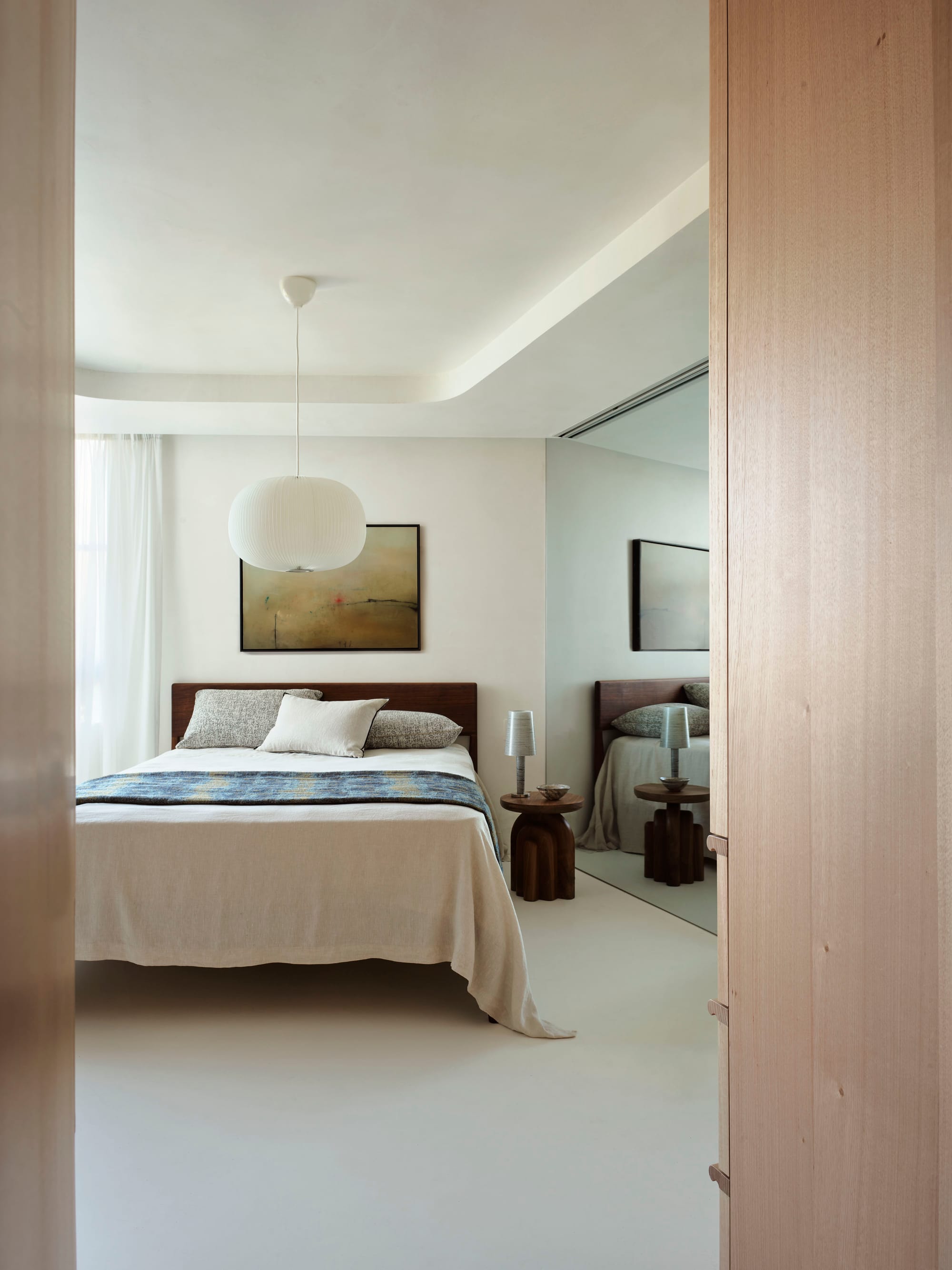 Tasmanian Timber Series: Darling Point Apartment. Photography by Ansen Smart. Vertical image of minimal bedroom in residential apartment. White neutral floors, walls and ceiling. Floor to mirror ceilings on the right hand side of the room. Curved white lamp hung above simple timber bedframe. 