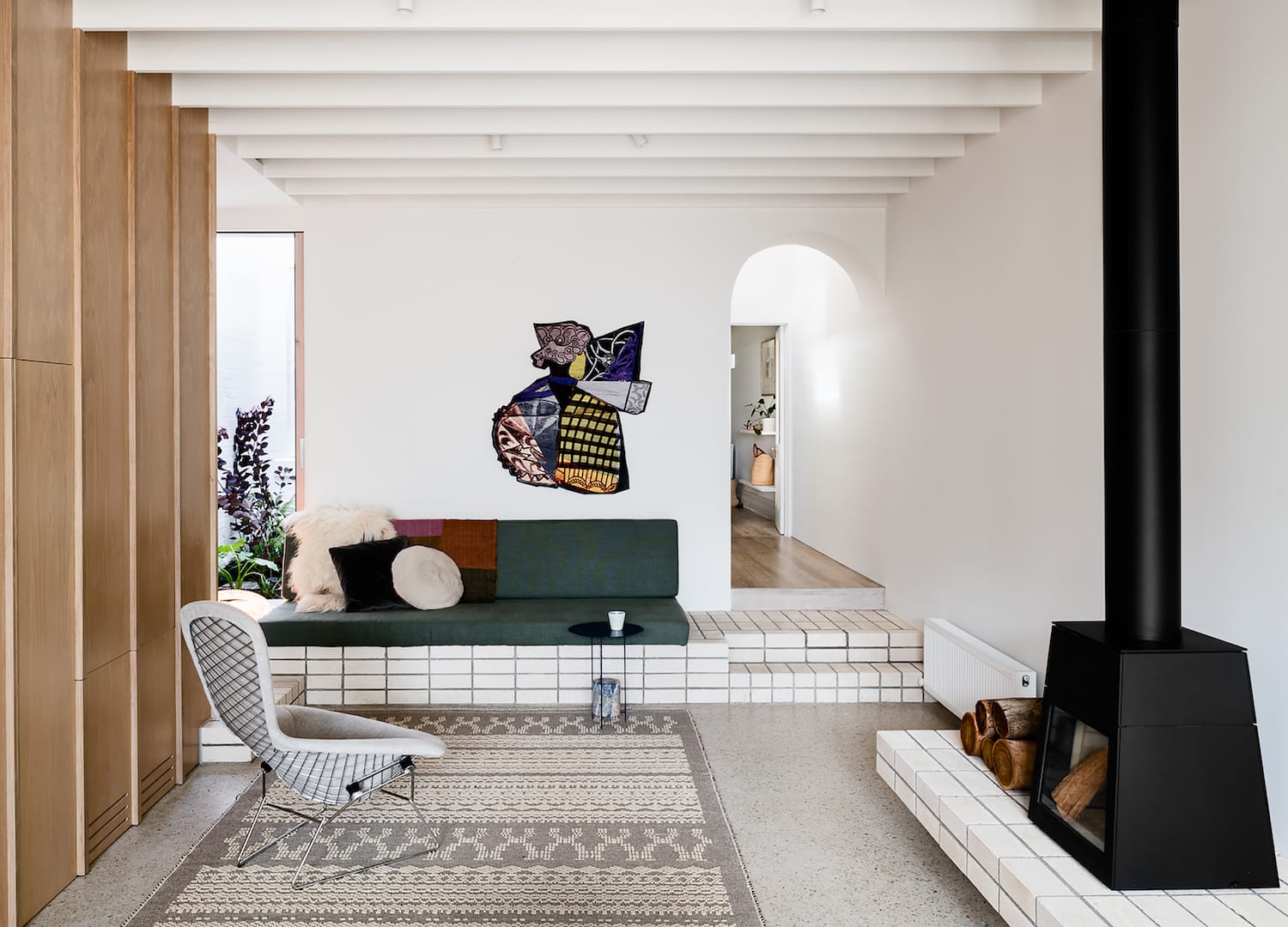 Storybook House by Folk Architects. Phtography by Tom Blachford. Landscape image of residential living space. Polished concrete floors and white walls. Curved archway in left background. Contemporary, black stand-alone fireplace left foreground. Integrated couch on elevated white brick surface. 