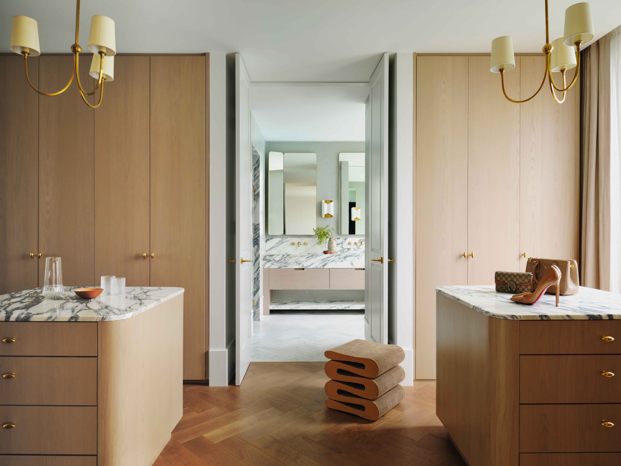 Light House by Smac Studio. Photography by Dave Wheeler. Landscape image of residential walk in wardrobe and ensuite. Herringbone floors, timber floor-to-ceiling cabinetry and islands. Marble countertops. White ceilings. Classic lightshades. White, airy marble bathroom in background.  