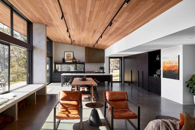 House Woodlands by AO Design Studio. Photography by Luc Remond. Wide angle shot of contemporary home. Polisged concrete floors. Timber panelled ceiling and black framed windows. Black modern kitchen. 