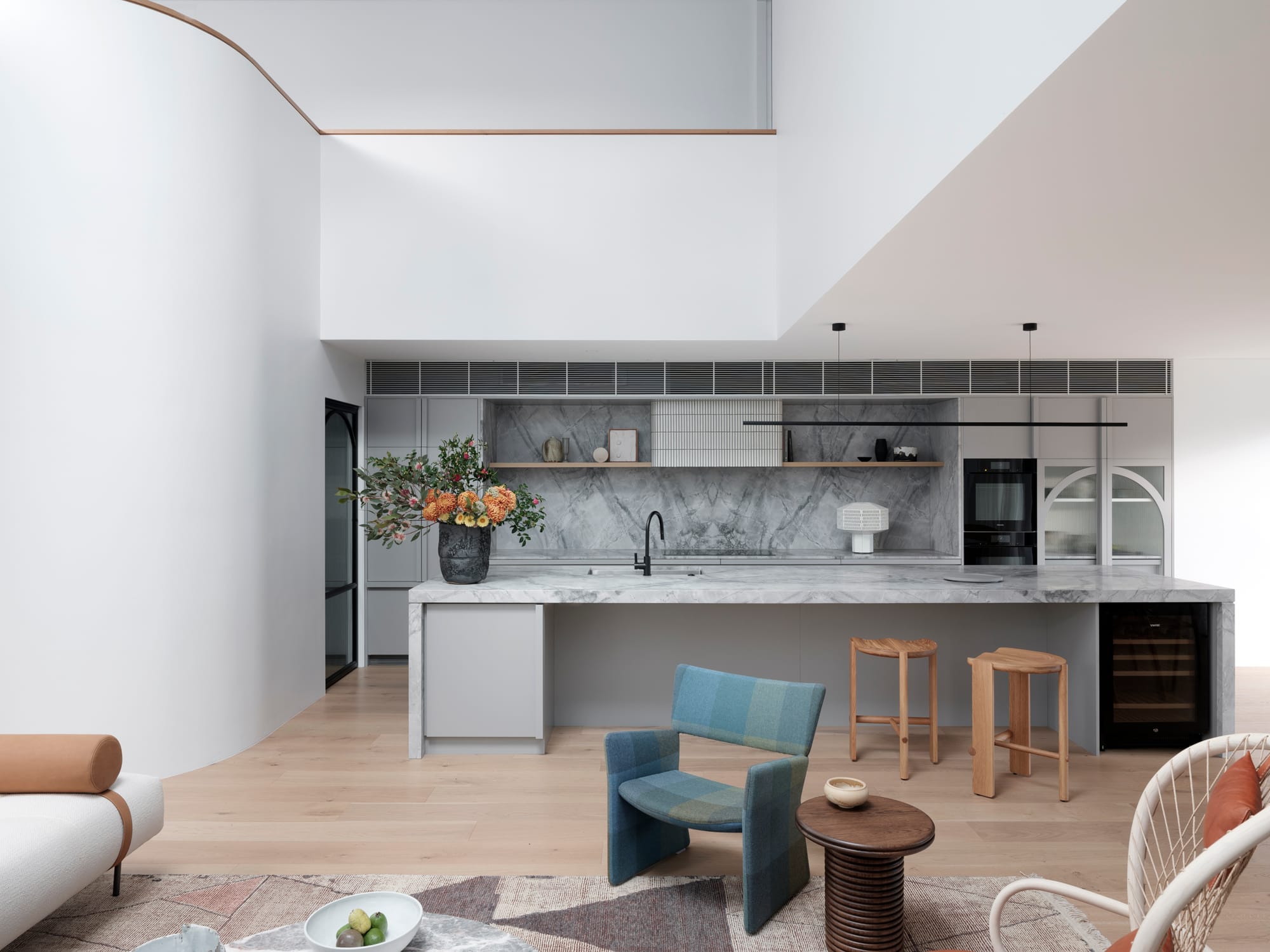 Bellevue House by Carla Middleton Architects. Photography by Tom Ferguson. Kitchen with marble and island benchtop. Assorted armchairs in living room in foreground. White walls and overhead void. 