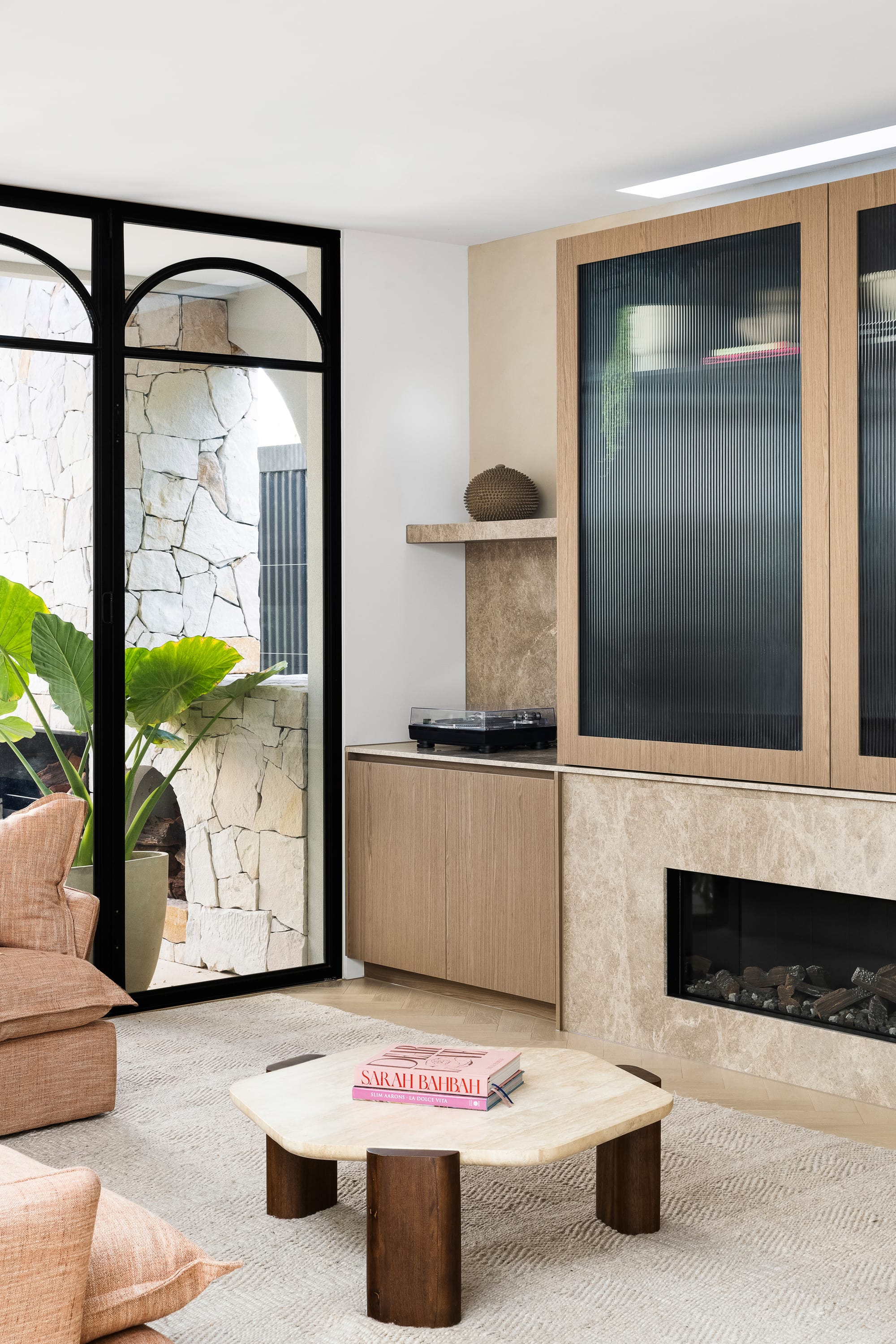 Goldsmith Residence by C.Kairouz Architects. Photography by Spacecraft. Living space with integrated timber cabinetry and timber and stone hexagonal coffee table. Tall black framed windows overlook outdoor courtyard. 