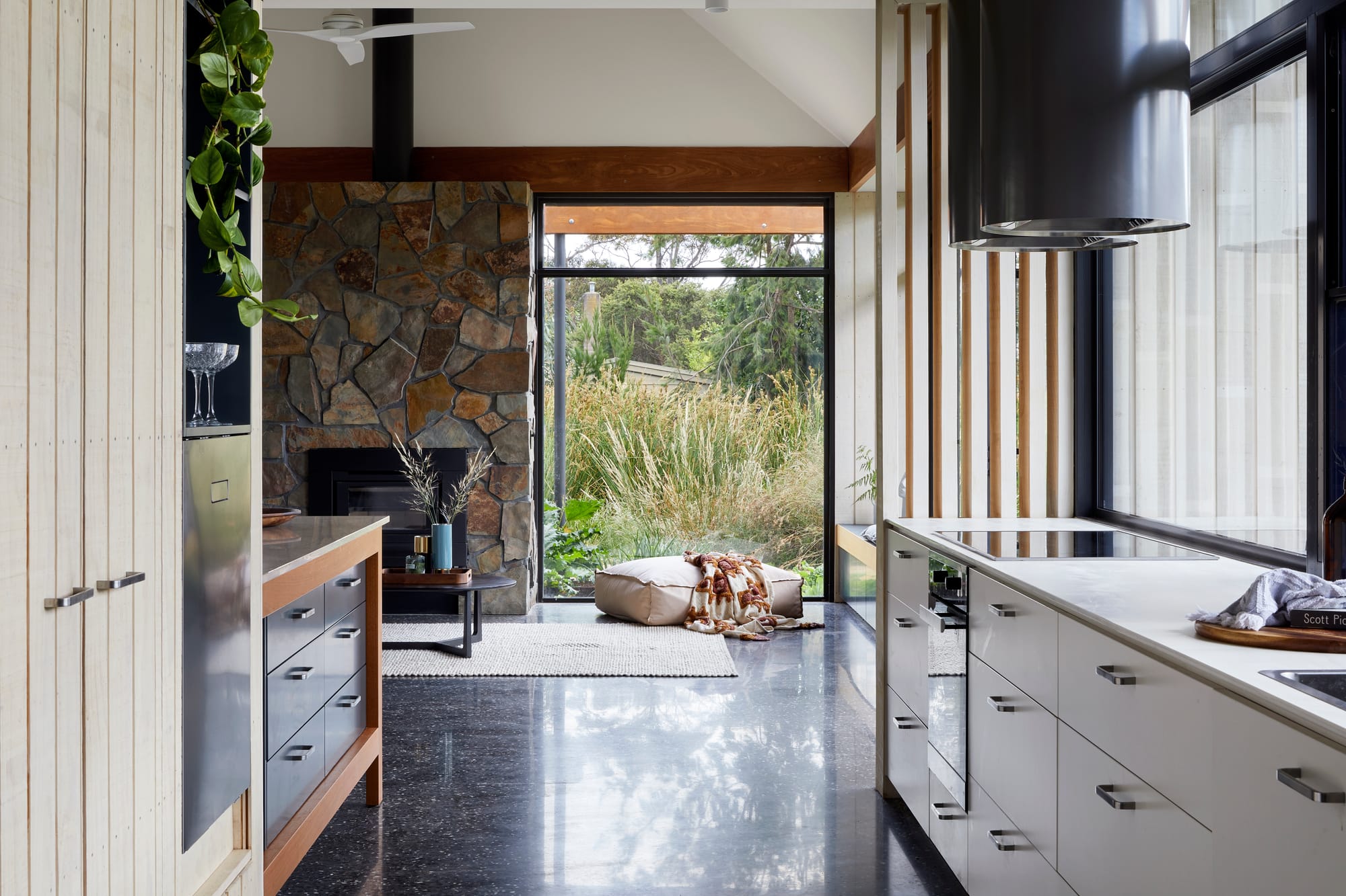 An interior shot of the kitchen with the living room in the background showing the large windows that help connect it to the landscape