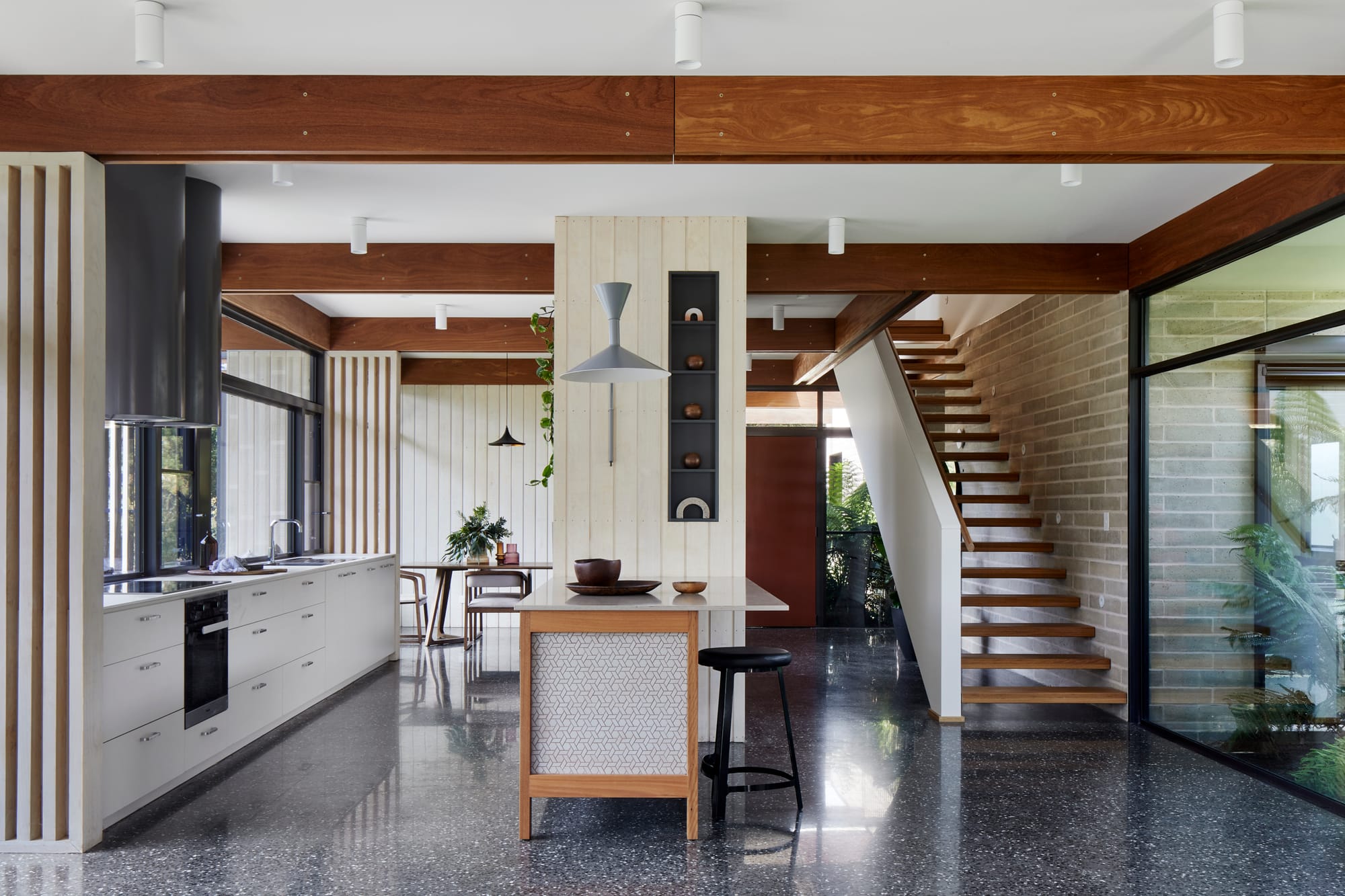 Mt Eliza House by BENT Architecture. Photography by Tatjana Plitt. Residential kitchen with island bench. Staircase to right of kitchen. Polished concrete floors and white clad walls. Exposed timber beams across ceiling. 