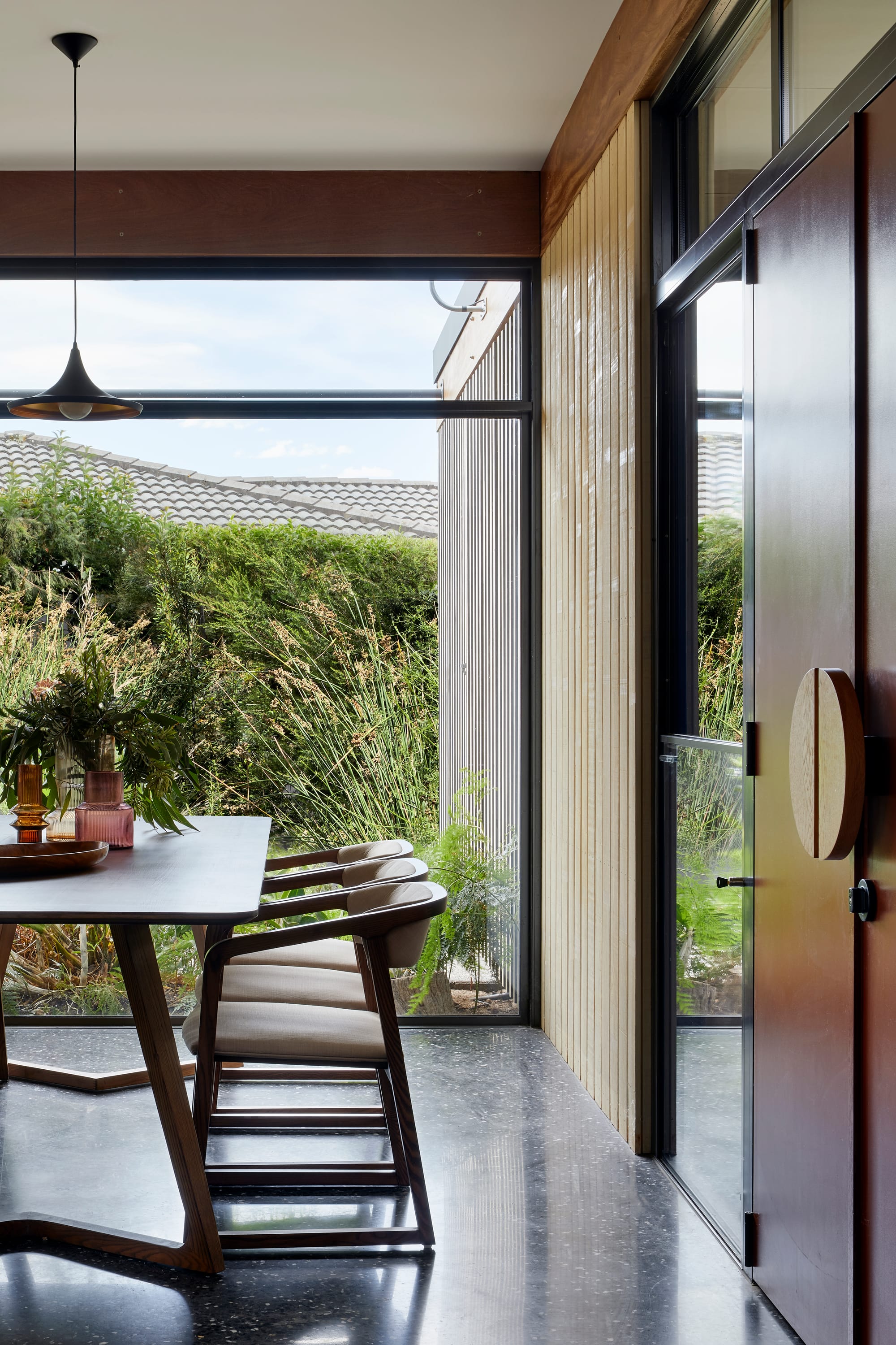 Mt Eliza House by BENT Architecture. Photography by Tatjana Plitt. Dining space with polished concrete floors and large windows overlooking exterior gardens. Large double doors to right of image and timber table and chairs to left. 