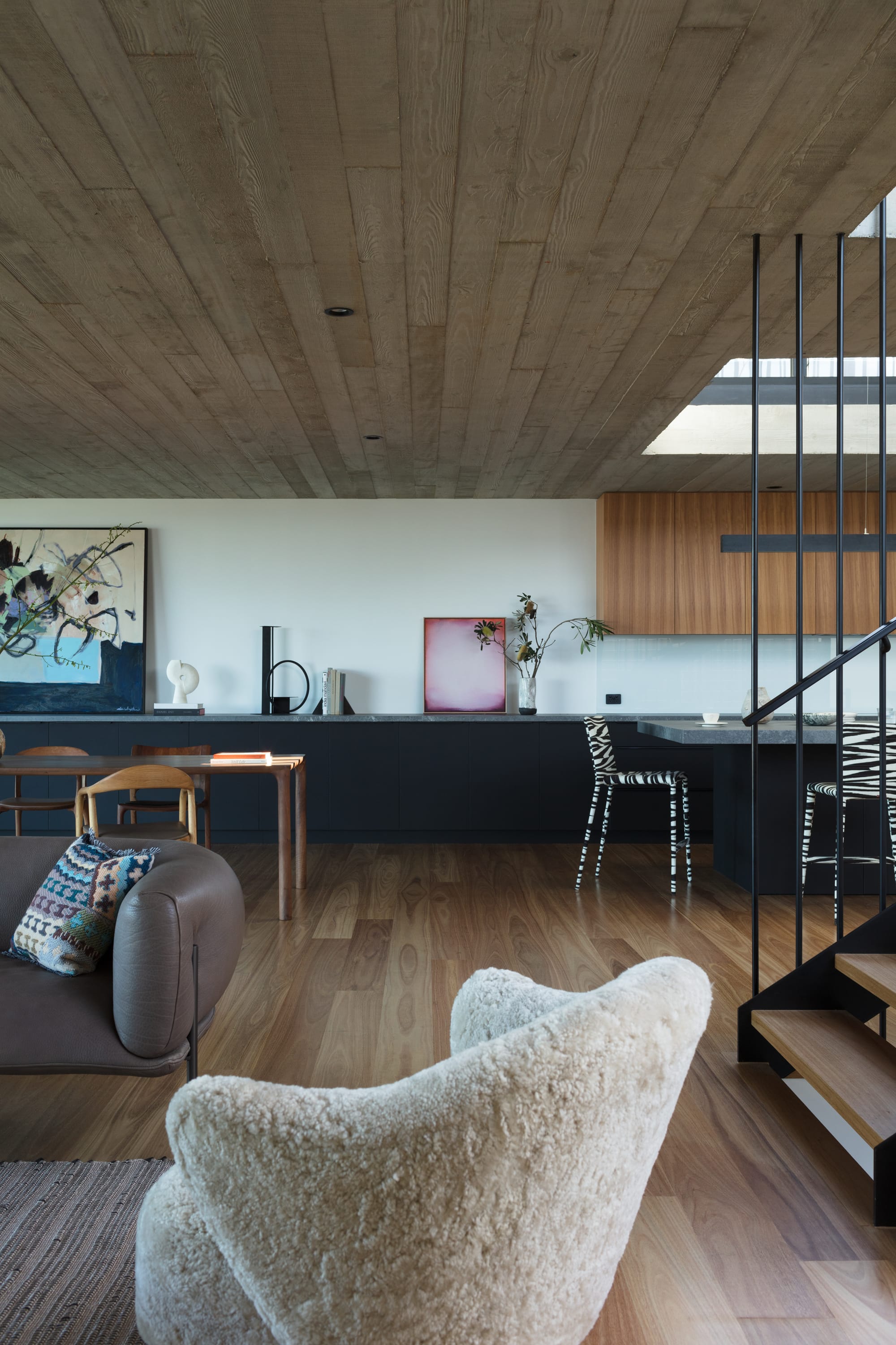 Grandview House by Ian Bennett Design Studio. Photography by Clinton Weaver. Open pan living, kitchen and dining area. Black cabinetry in background. White fluffy armchair in foreground. Timber floors throughout. 
