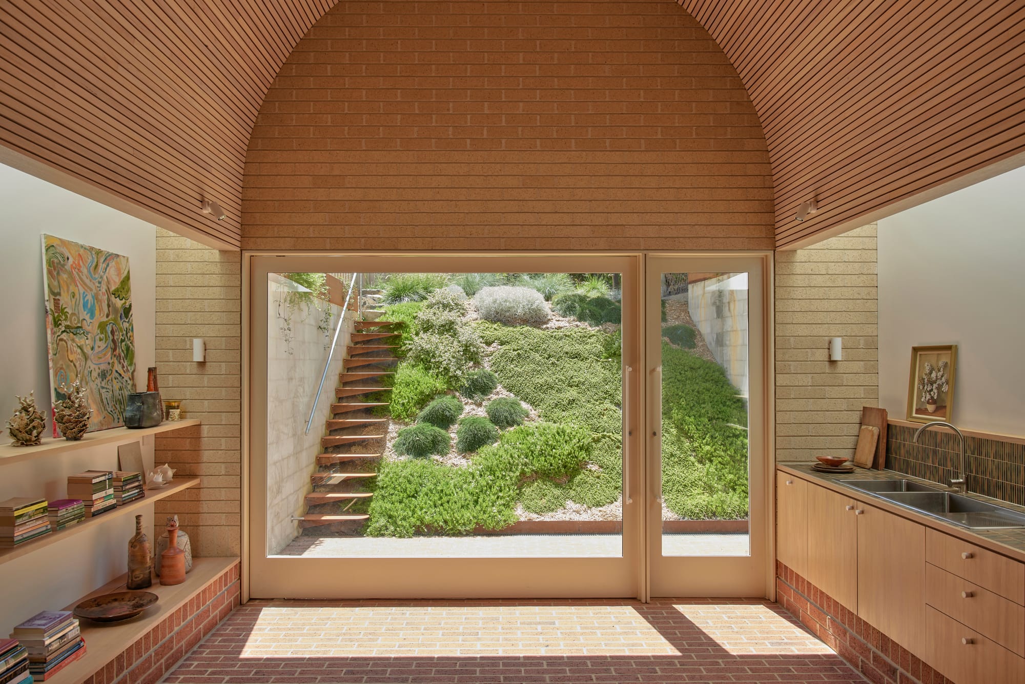 Harriet's House by SO:Architecture. Photography by Sean Fennessy. Landscape shot of open plan kitchen area with high arched ceiling and brick floors. Windowed wall overlooks sloping garden. 