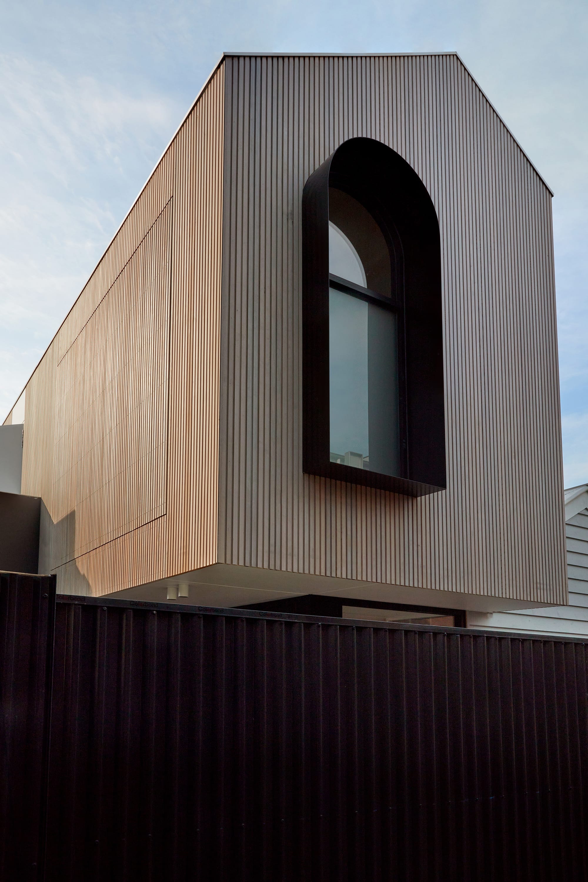 The Full Monty by FYC Architects. Photography by Stephanie Rooney. Exterior facade of residential home with tall gabled roof. Timber clad. Black metal framed window and fence. 