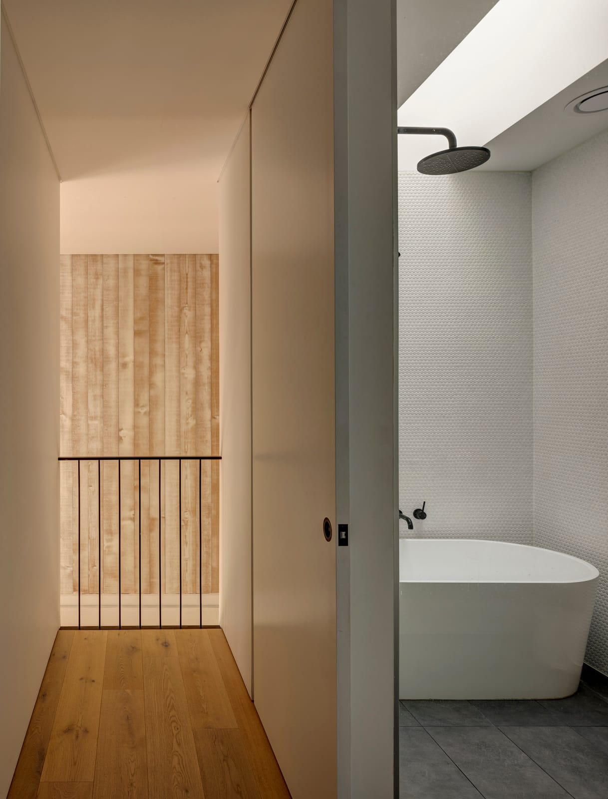 Quarry Box by MCK Architects. Photography by Brett Boardman. Hallway with timber floors next to a white and grey tiled bathroom to the right. 