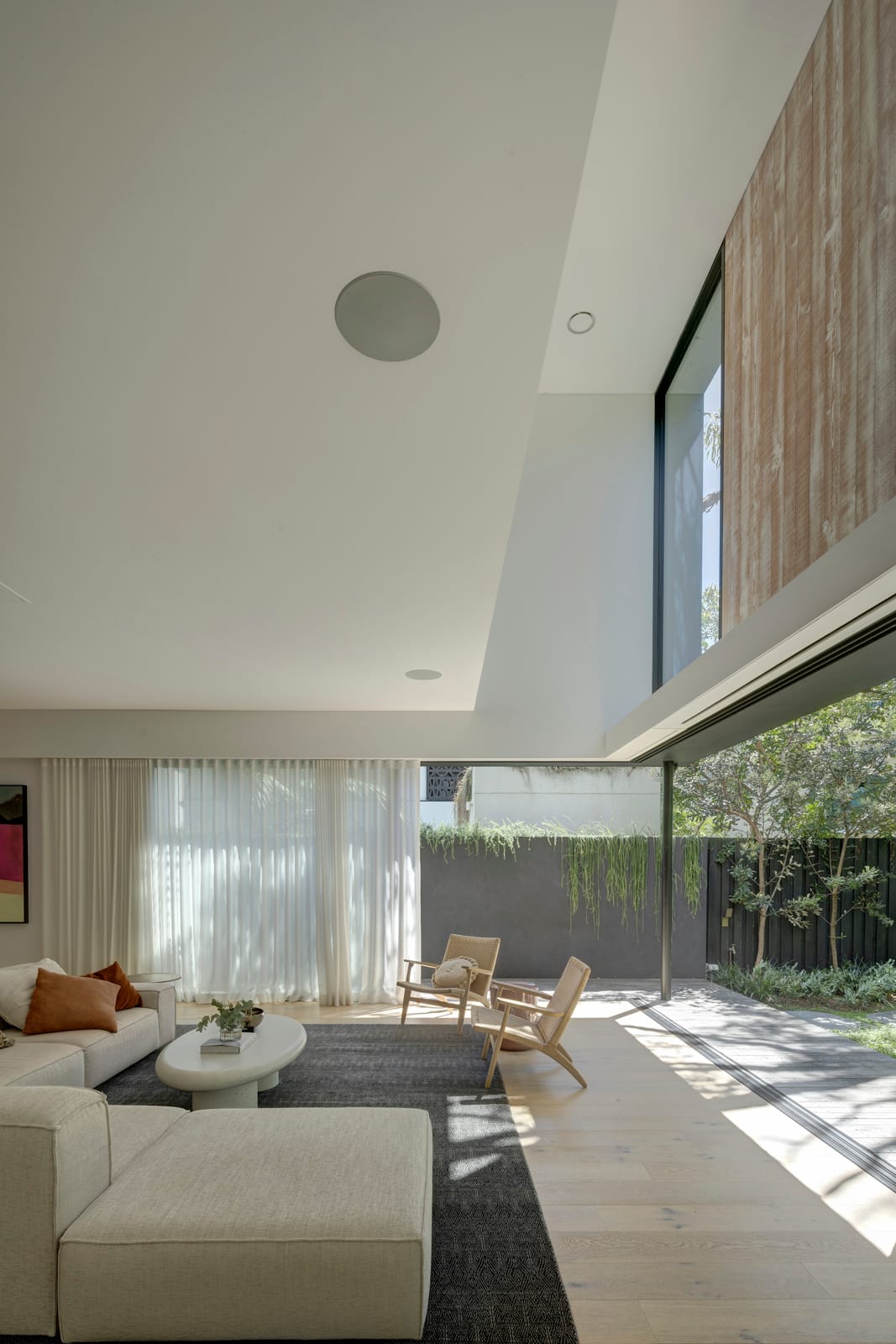 Quarry Box by MCK Architects. Photography by Brett Boardman. Living room with pale timber floors, large grey rug and oat-coloured couch. Floor-to-ceiling windows overlook grey fence. 