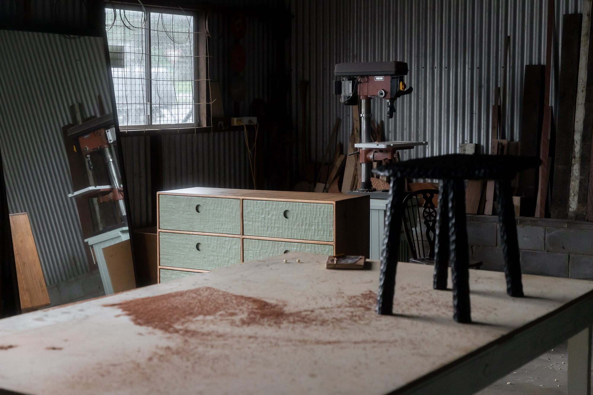 Two Blue Boys. Copyright of Two Blue Boys. Inside of workshop, with timber and seafoam sideboard pictured in background. Small black timber stool on construction table.