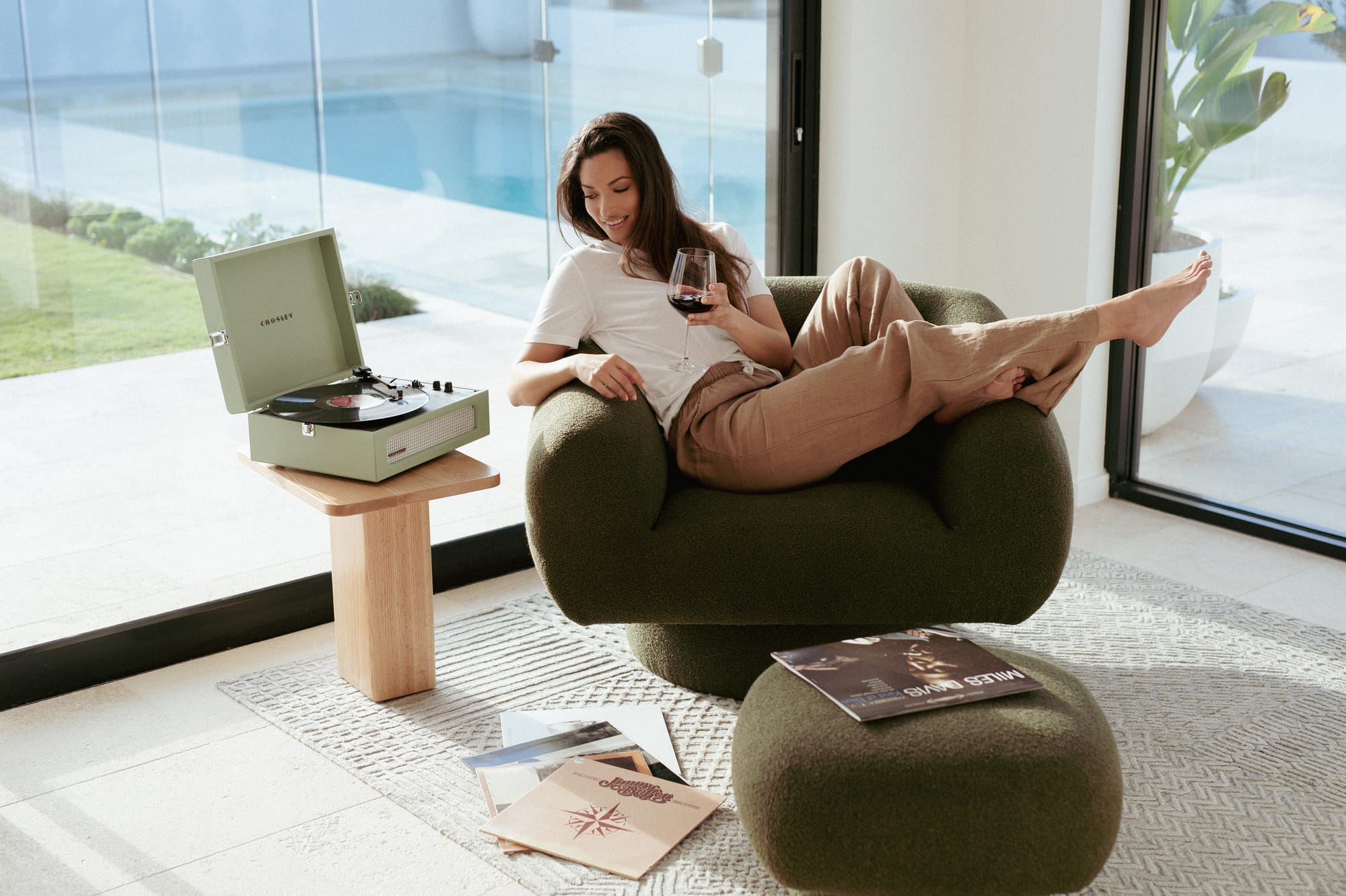 Made Furniture. Copyright of Made Furniture. Woman lounging in olive armchair with wineglass. Record player on stool next to chair, footstool on rug. 