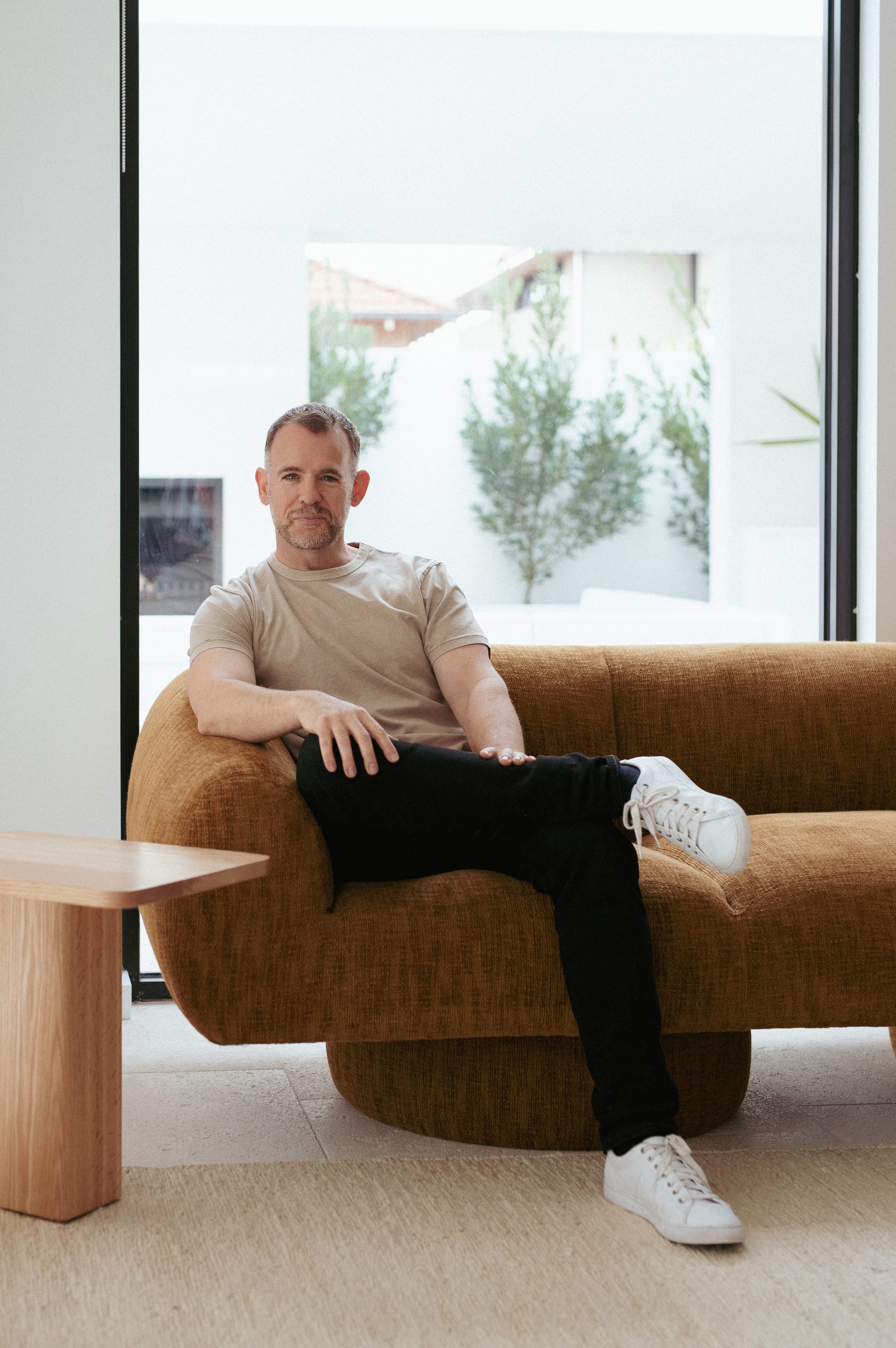 Made Furniture. Copyright of Made Furniture. Man sitting in burnt orange sofa. Timber side table to the left. Large window behind couch. 