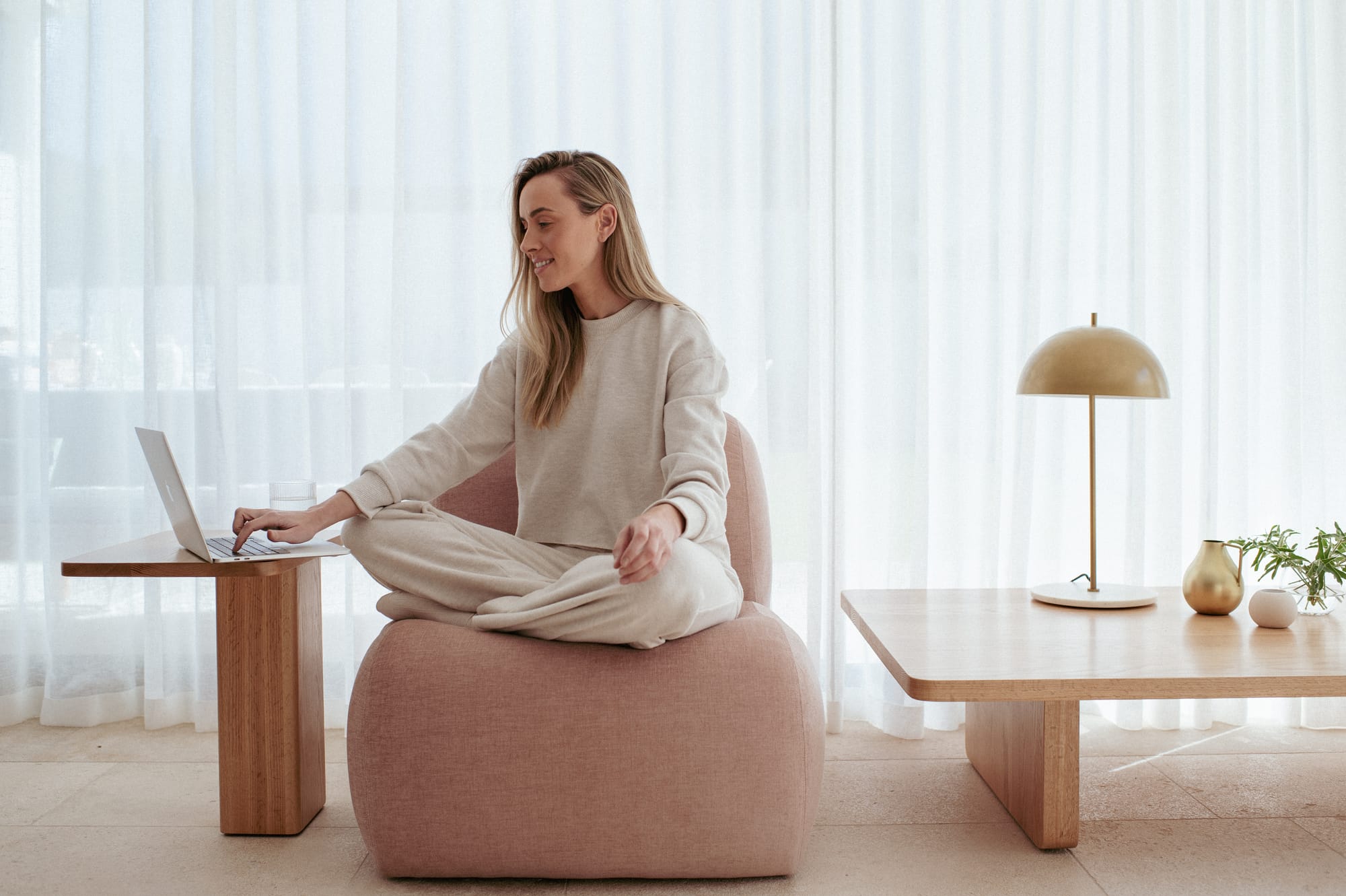 Made Furniture. Copyright of Made Furniture. Woman sitting on rose coloured armchair with timber side table to the left and timber coffee table to the right. 