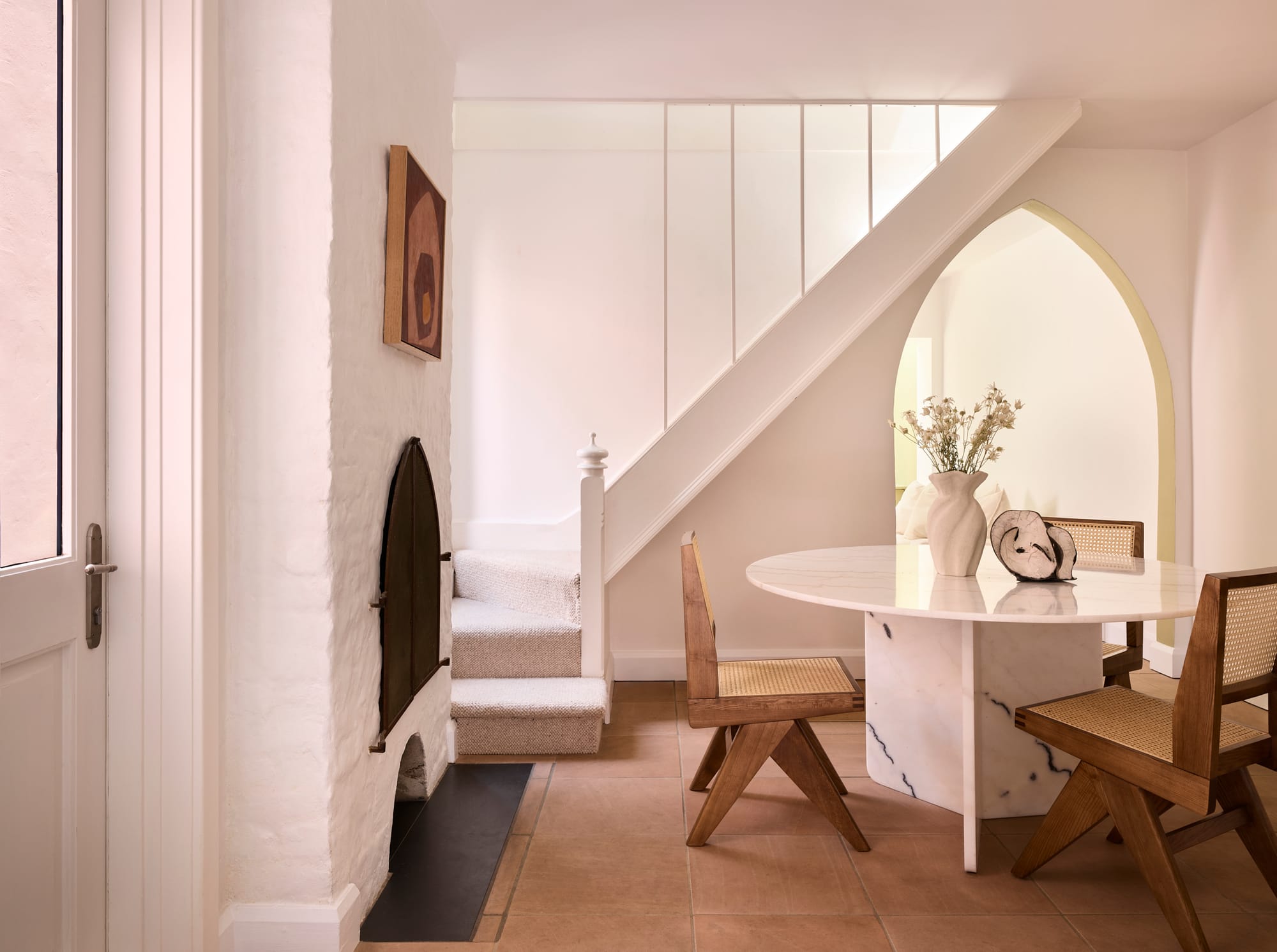 Chapel House by FURNISHD. Photography by Alicia Taylor. Round marble dining table with ratan mid-century chairs on tiled floor. White brick fireplace and white staircase.