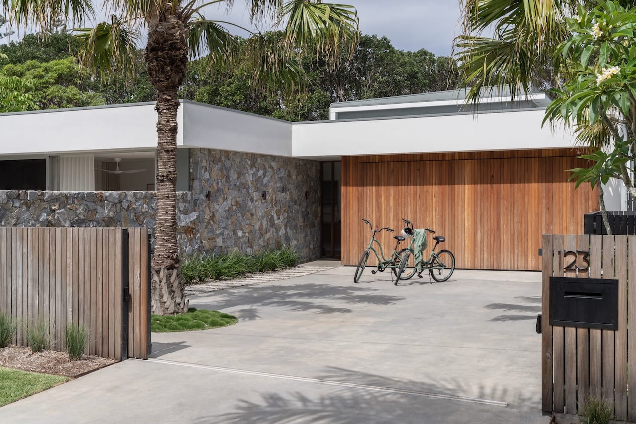 Kasa Byron Bay. Photography by Adam Nicolson. Street facade of home with timber garage door, stone wall and concrete driveway. Two bikes sit on driveway. 