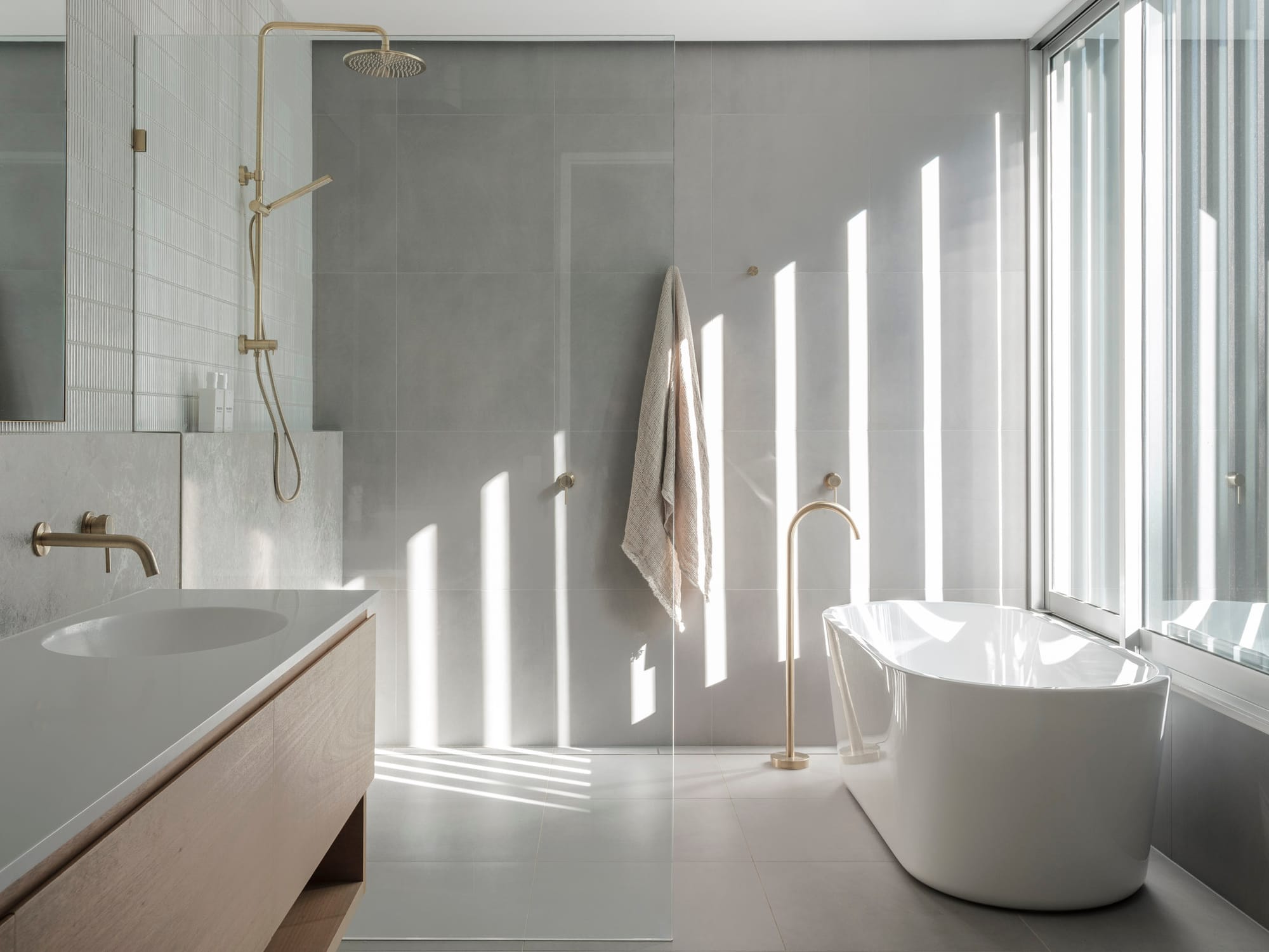 Kasa Byron Bay. Photography by Tom Ferguson. Bathroom with grey tiled floors and walls. Freestanding white tub to right. Gold tapware. Timber vanity to left. 