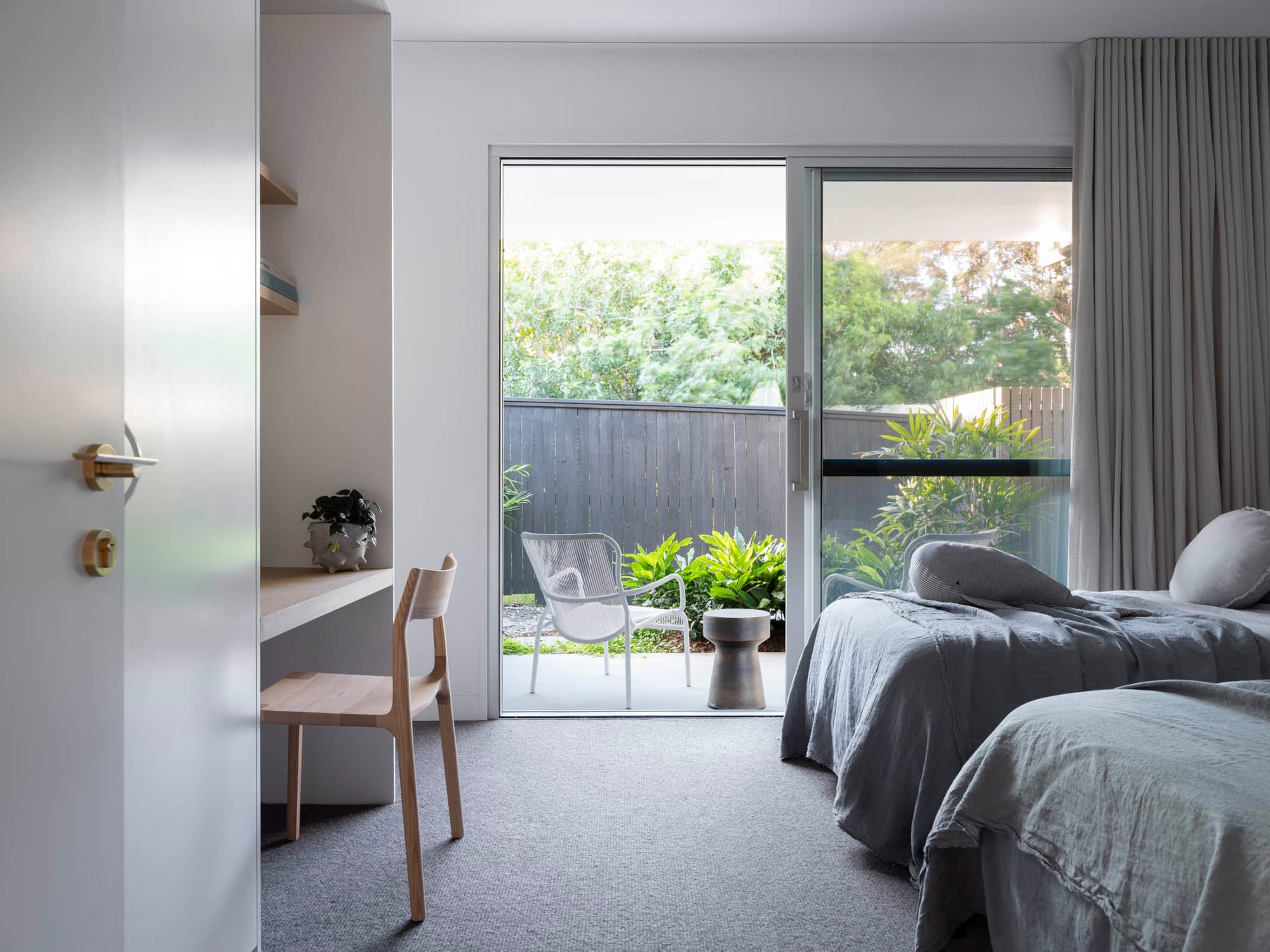 Kasa Byron Bay. Photography by Tom Ferguson. Bedroom opening onto courtyard with chair and gardens. Grey carpets and two single beds inside. Timber chair at study nook to left. 