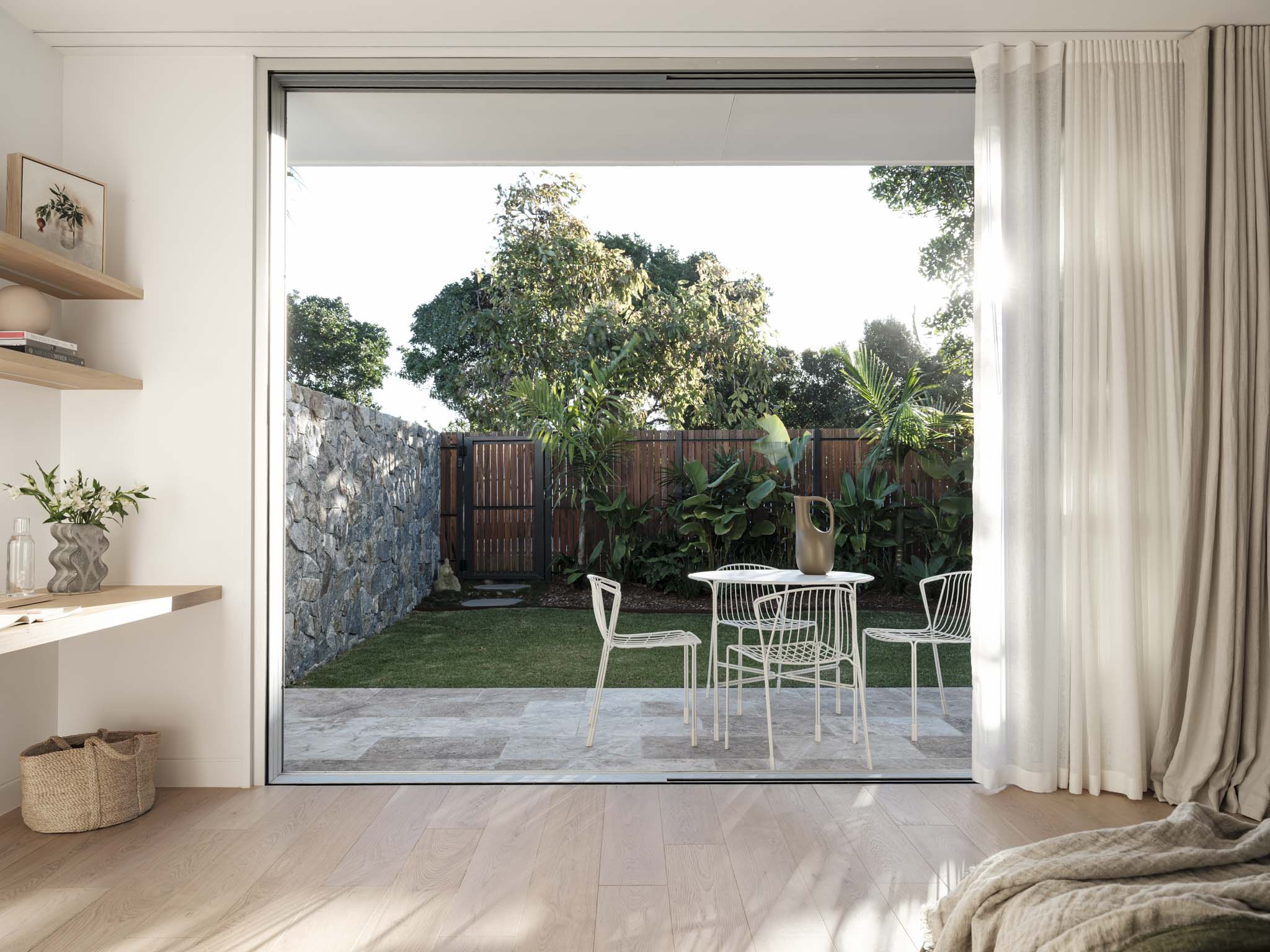Kasa Byron Bay. Photography by Tom Ferguson. Floor-to-ceiling door opening onto tiled patio and garden with dining setting. Timber floors and white walls inside. 