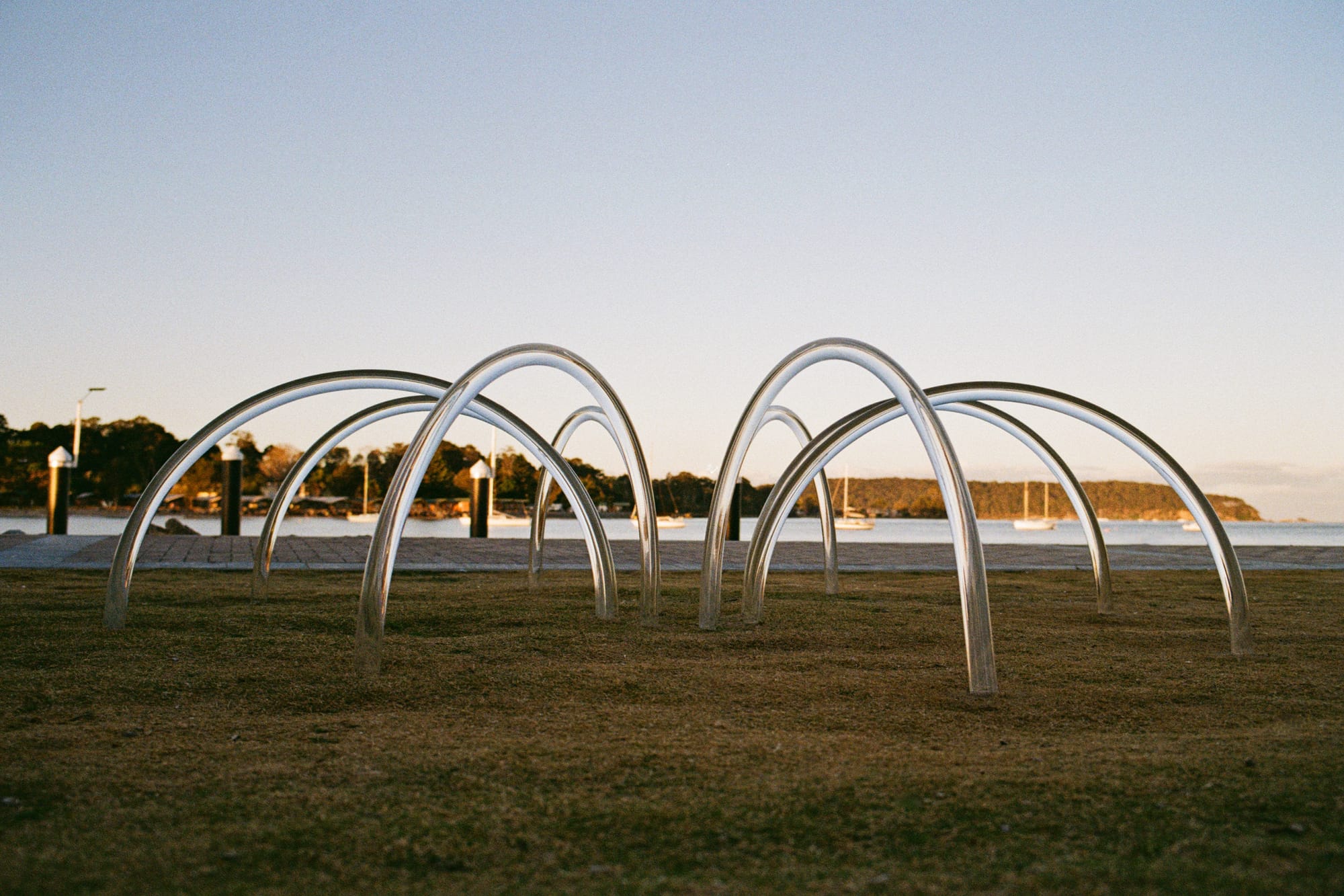 Array 16 by Studio Shand. Photography by Benjamin Jay Shand. Abstract installation art constructed of clear, curved material. Sculpture on grass in front of jetty and body of water. 