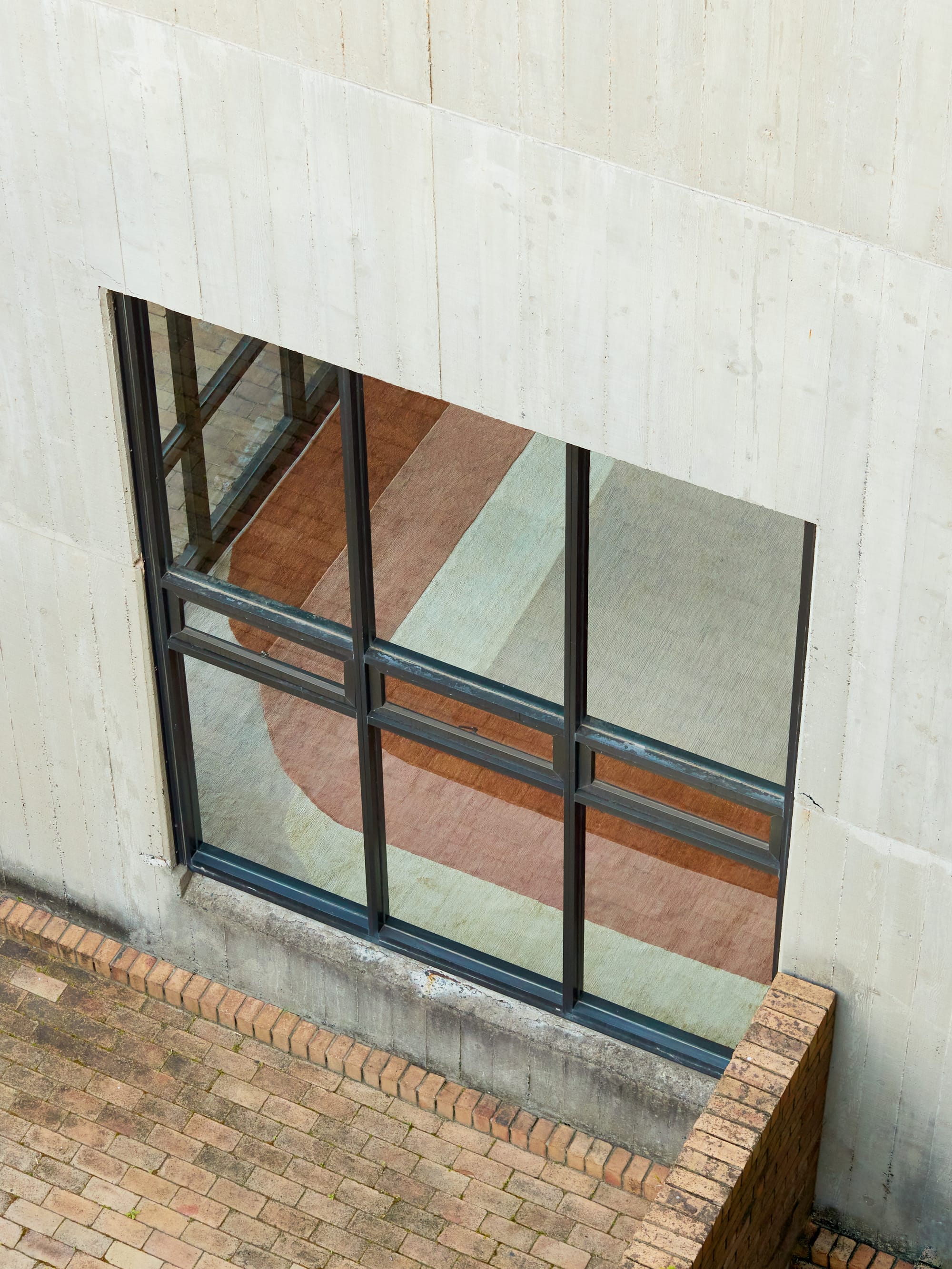 SUPERGRAPHICS Collection for Cadrys by Studio Shand. Photography by Traianos Pakioufakis. Black framed window on white wall, taken from extreme high angle. Whitw, red and orange abstract rug on floor visible through window. Brick floor exterior.