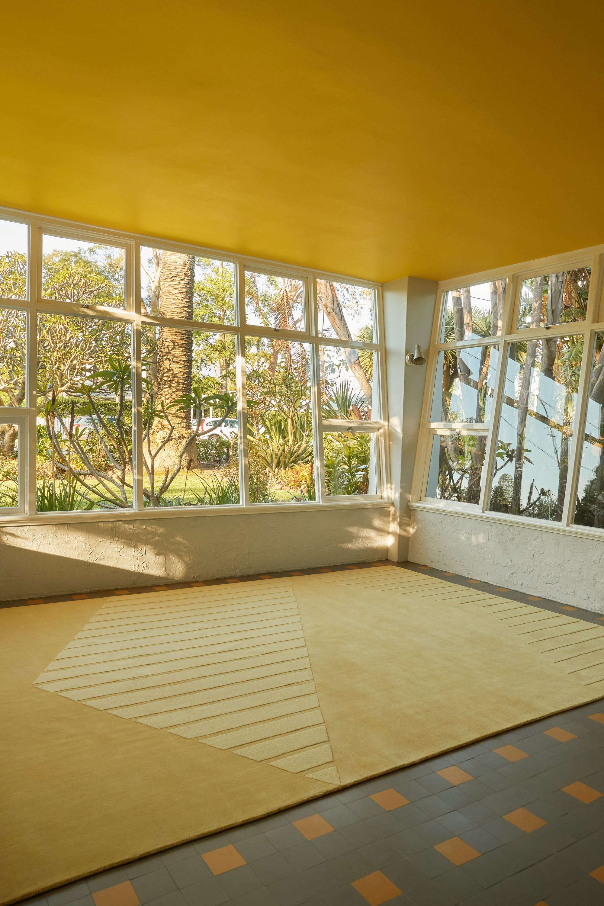 SUPERNOVA Collection for Cadrys by Studio Shand. Photography by Traianos Pakioufakis. Large yellow rug on tiled floor. Large, windowed walls overlooking lush suburban garden.