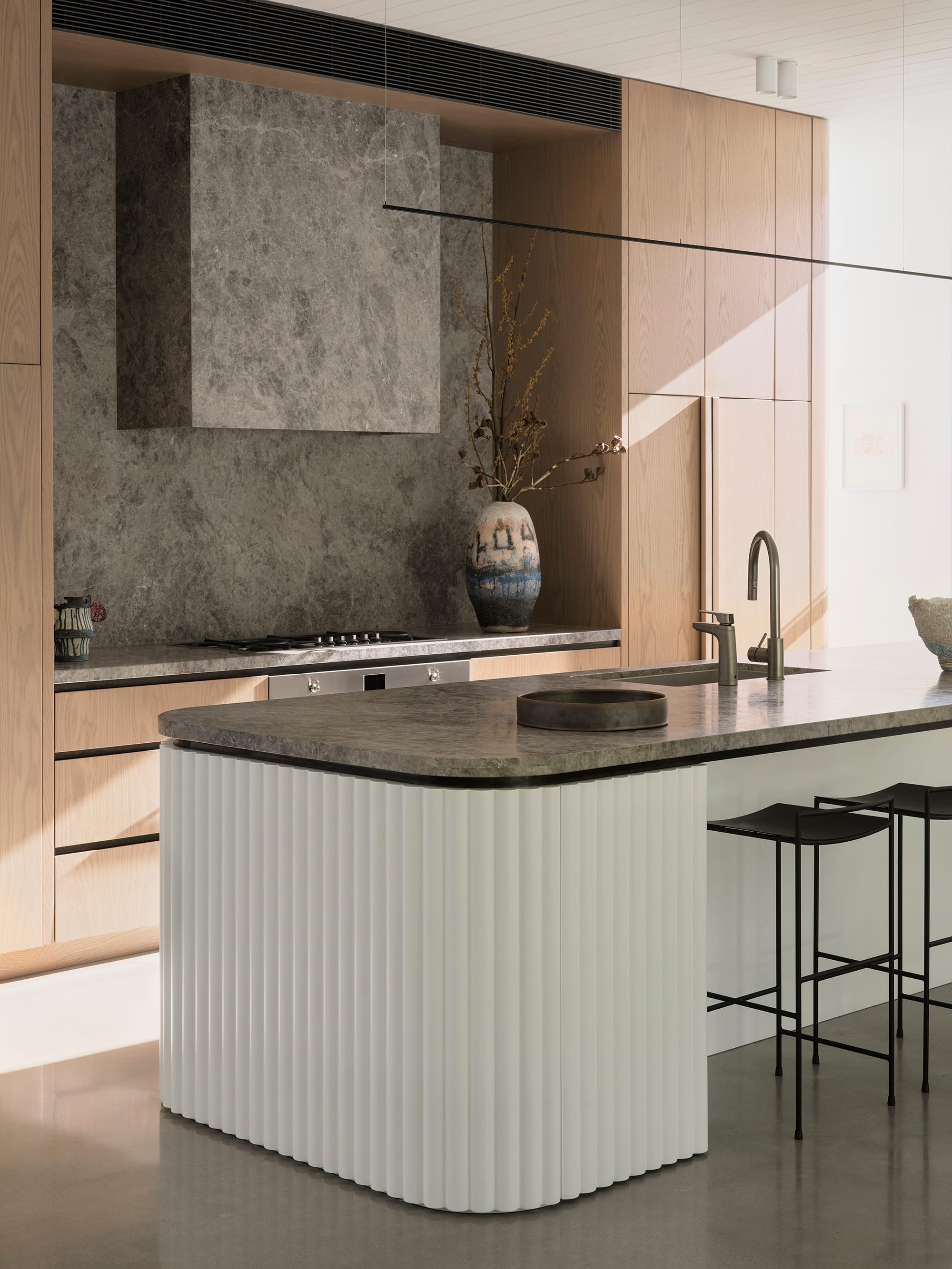 Warren House by CM Studio. Photography by Nic Gossage. Kitchen with white fluted island bench with stone countertop. Polished concrete floor. Timber kitchen cabinetry. 