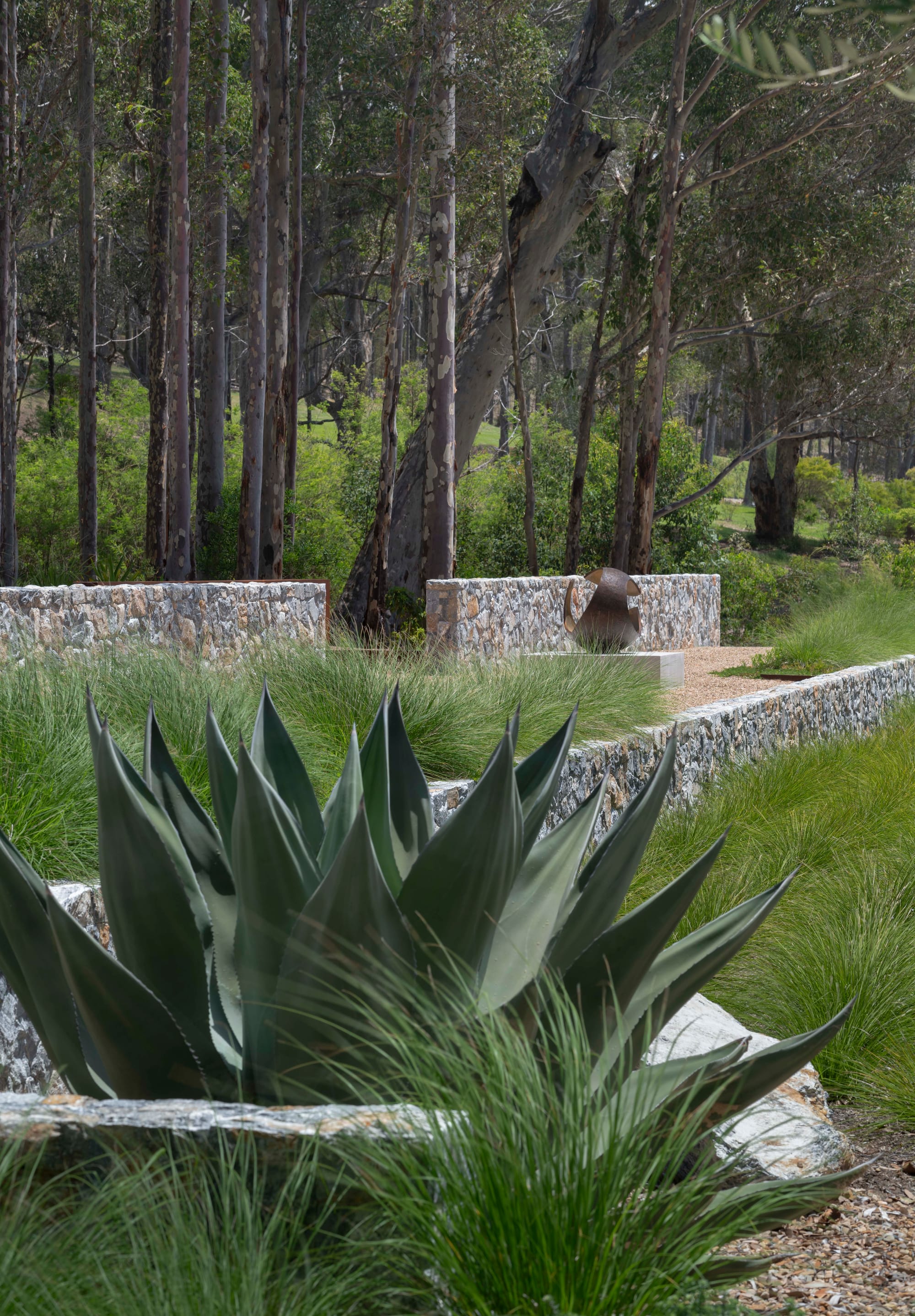 Bracken by Secret Gardens. Photography by Nicholas Watt. Agave plant in foreground. Stone retaining walls in garden in background. Spherical metal sculpture. 