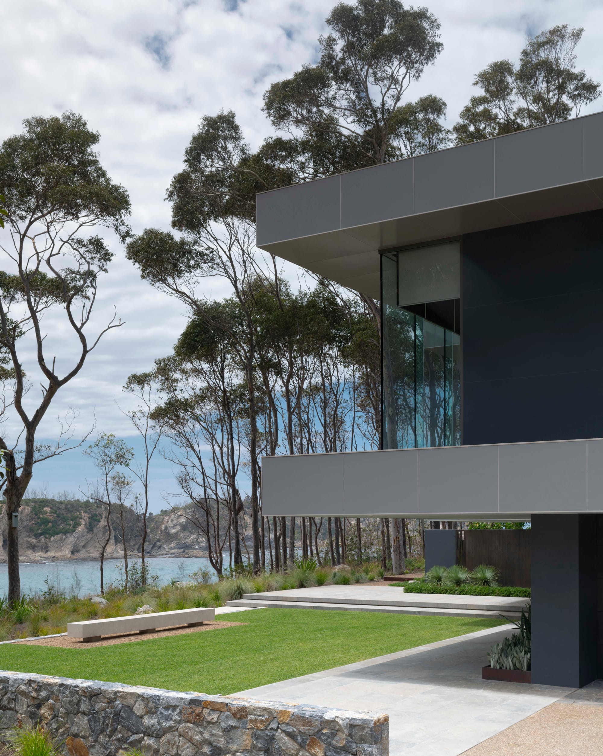Bracken by Secret Gardens. Photography by Nicholas Watt. Contemporary double storey home to the right of image. Neat green lawn overlooking rocky coastline to the left. Stone walls and low concrete paths.