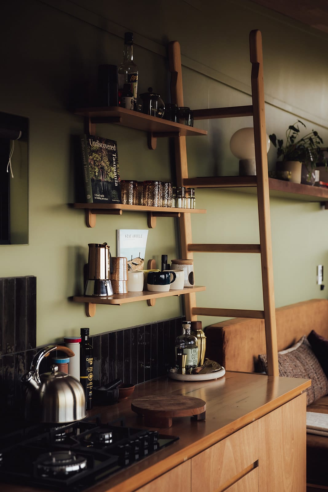 Candlebark Ridge. Copyright of Candlebark Ridge. Timber kitchen bench with stovetop and display shelving. Timber ladder leans against shelving. Black splashback tiles on sage green wall. 