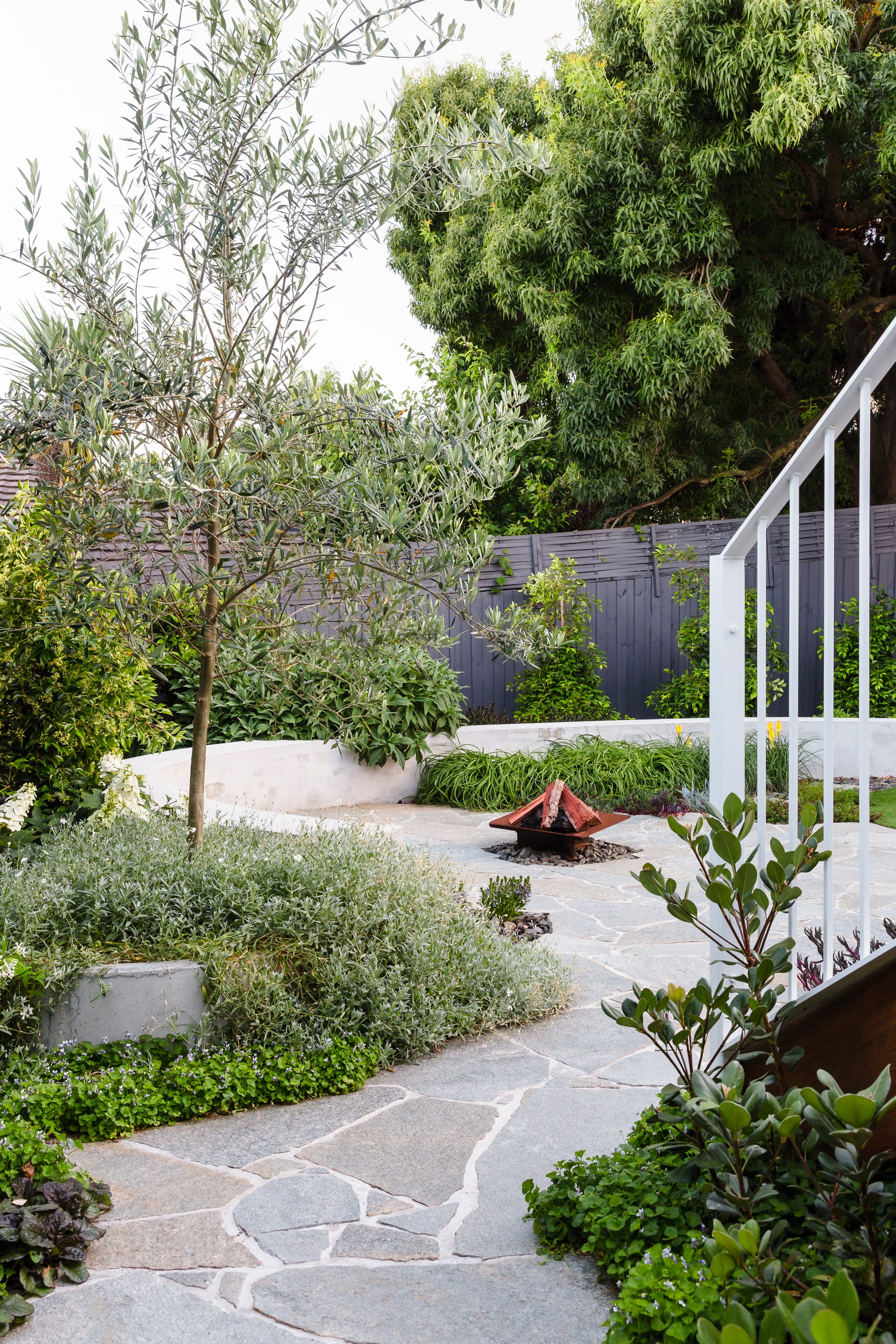 Hope Preston by Julie Crowe Landscape Design. Photography by Erik Holt Photography. Outdoor firepit with circular paved flooring and curved bench seating. Shrubs and trees grow around bench seating. 