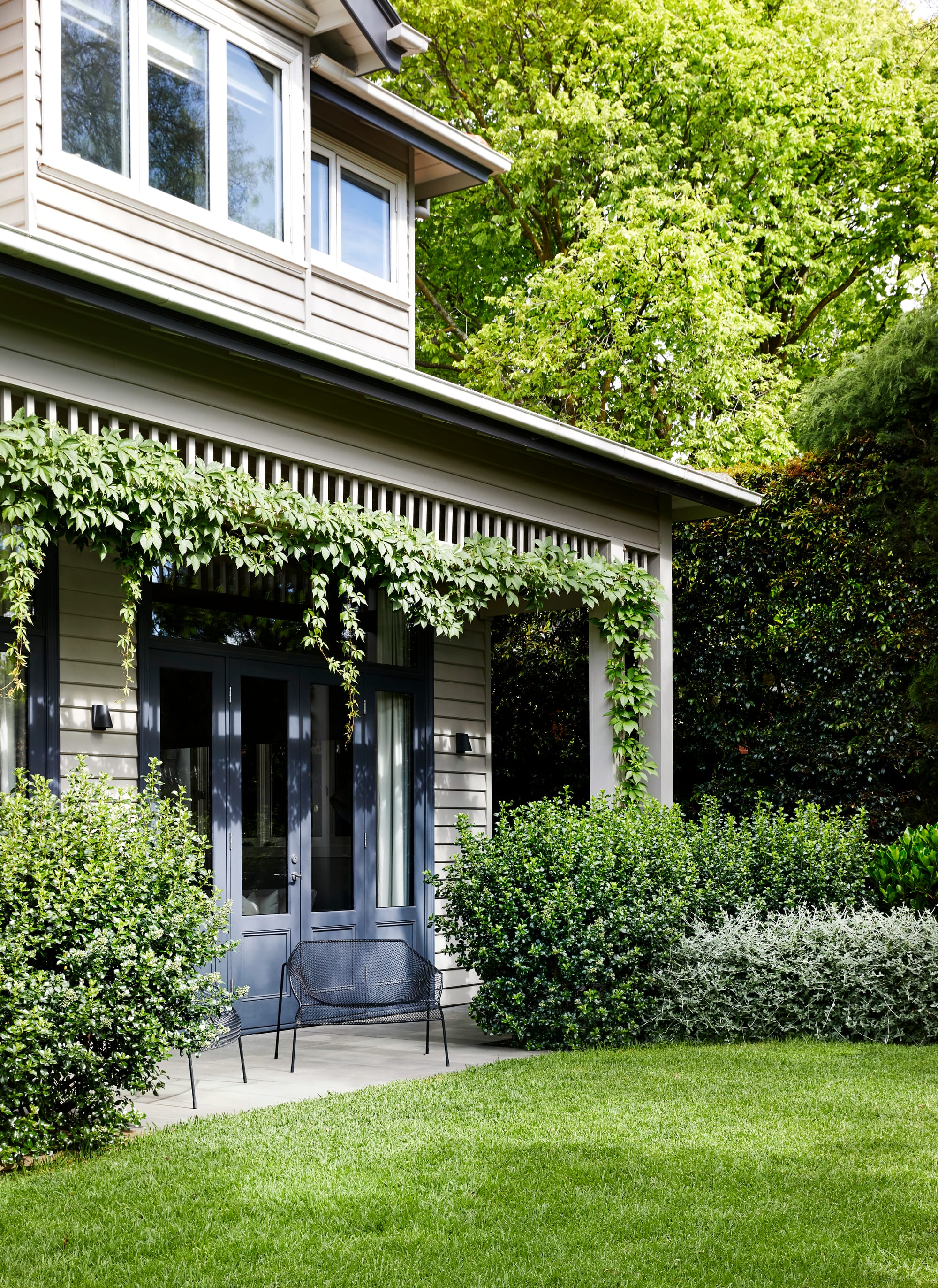 Julie Crowe Landscape Design.Photography by Caitlin Mills Photography. Double storey clad home with plants growing across verandah awnings. Green lawn and lush garden beds surround home.
