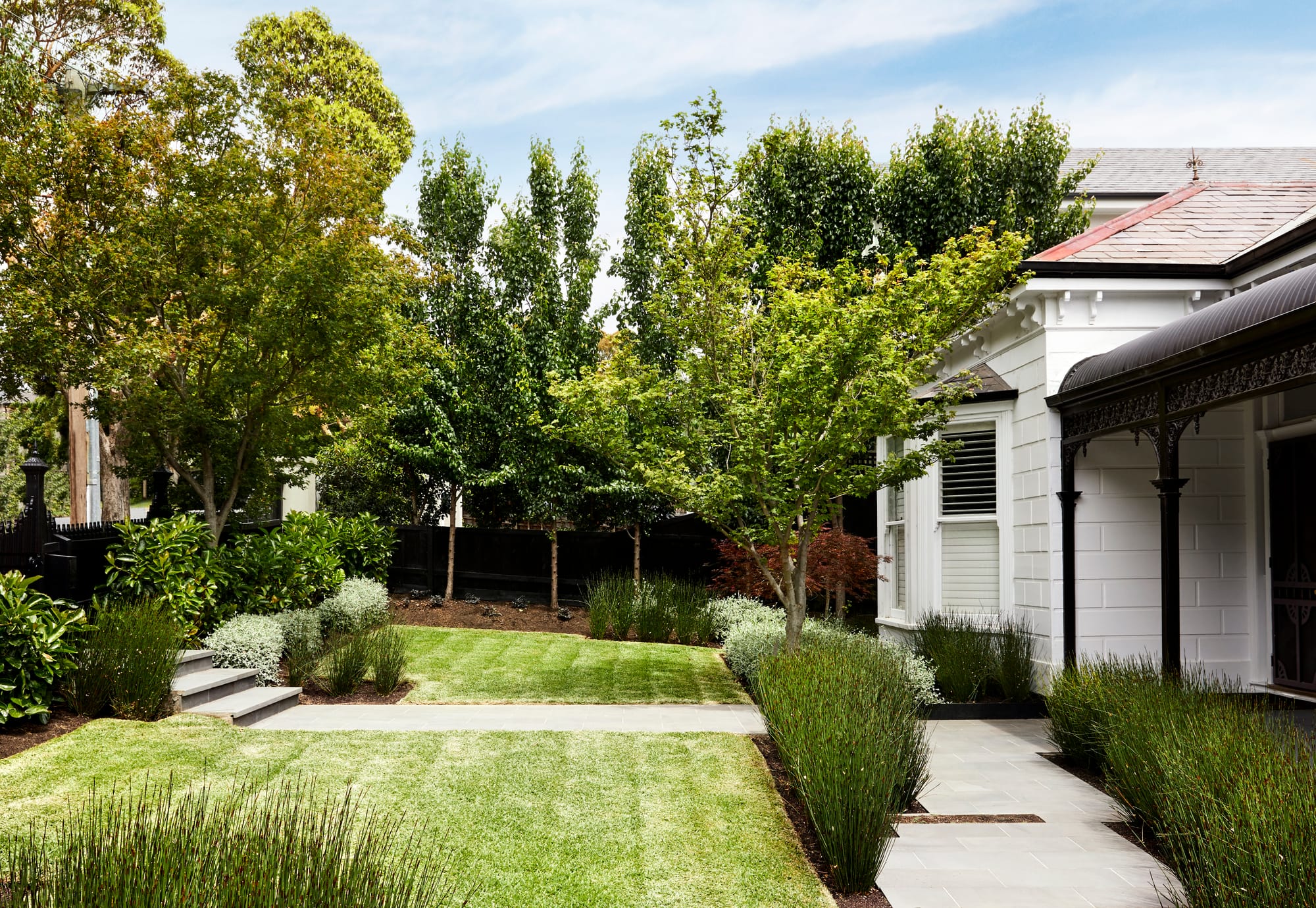 Julie Crowe Landscape Design. Photography by Caitlin Mills Photography. Multi-level garden extending from white brick Victorian-era home. Path cuts through green grass up to steps. Tall green trees dotted through garden beds.