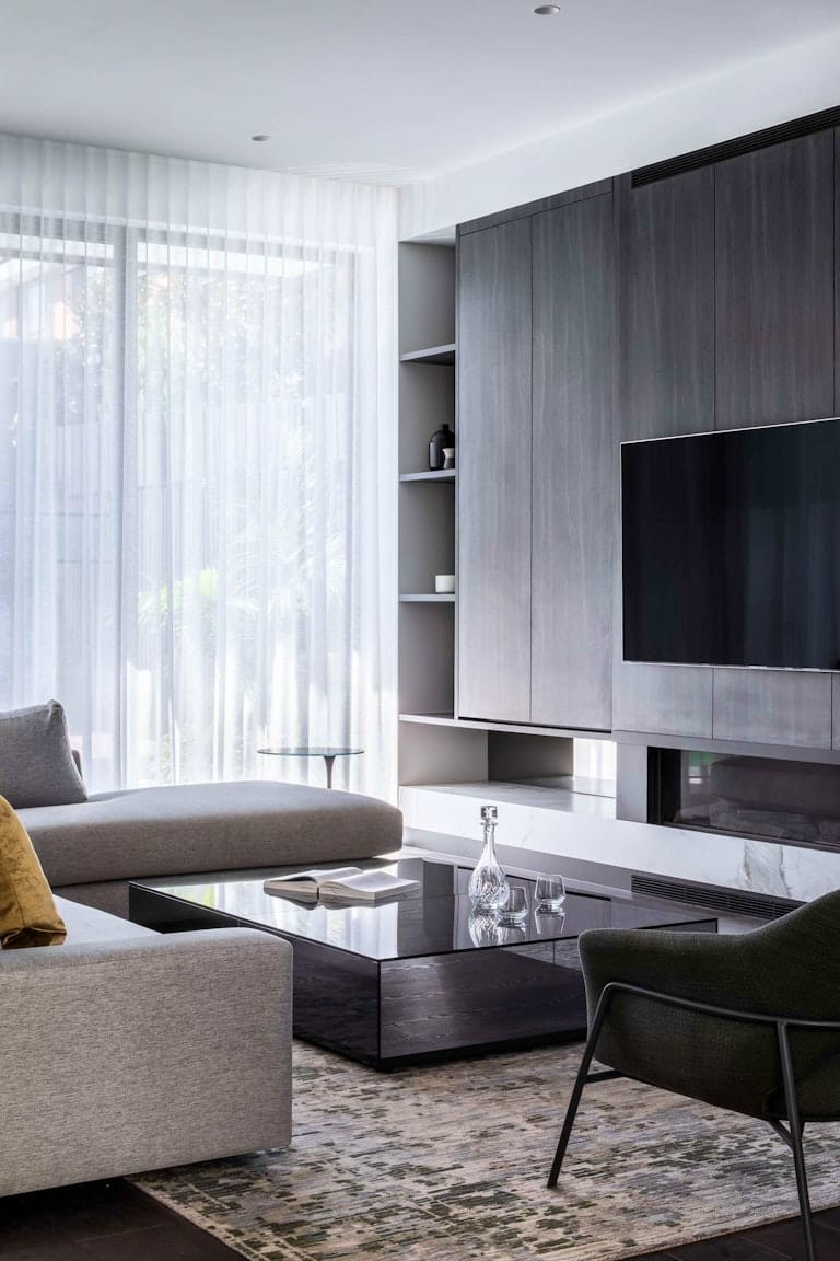 Northcote Renovation by Chan Architecture. Photography by Tatjana Plitt. Contemporary living space with grey timber cabinetry, dark timber floors and grey furniture. Sheer white curtains hang in front of floor-to-ceiling windows in background. 