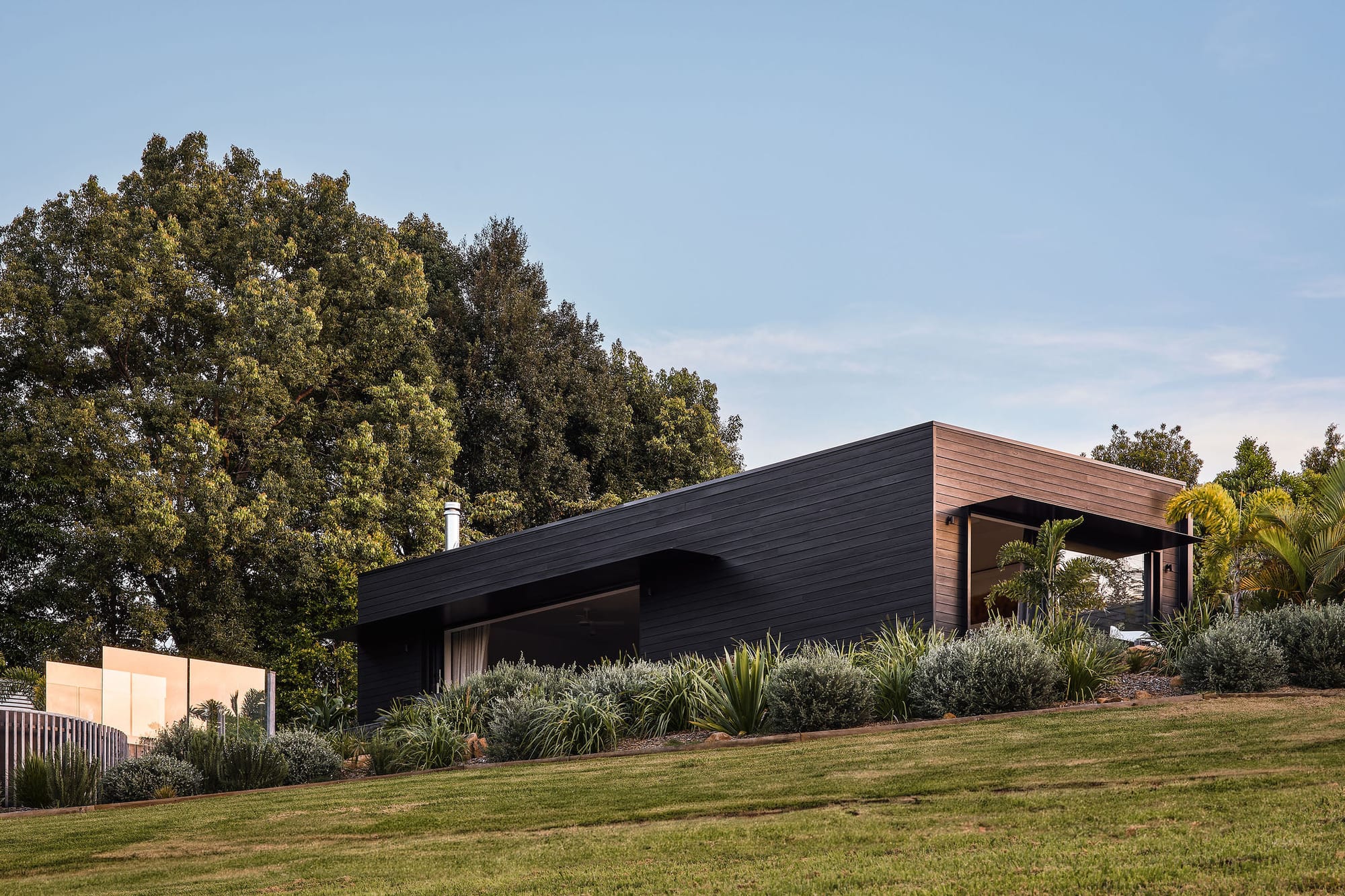 Rockpool Farm Byron Bay. Photography by Andy Macpherson. Rectangular timber clad building on top of grassy hill. Blue sky. Lots of plant life in garden bed in front of building and around.