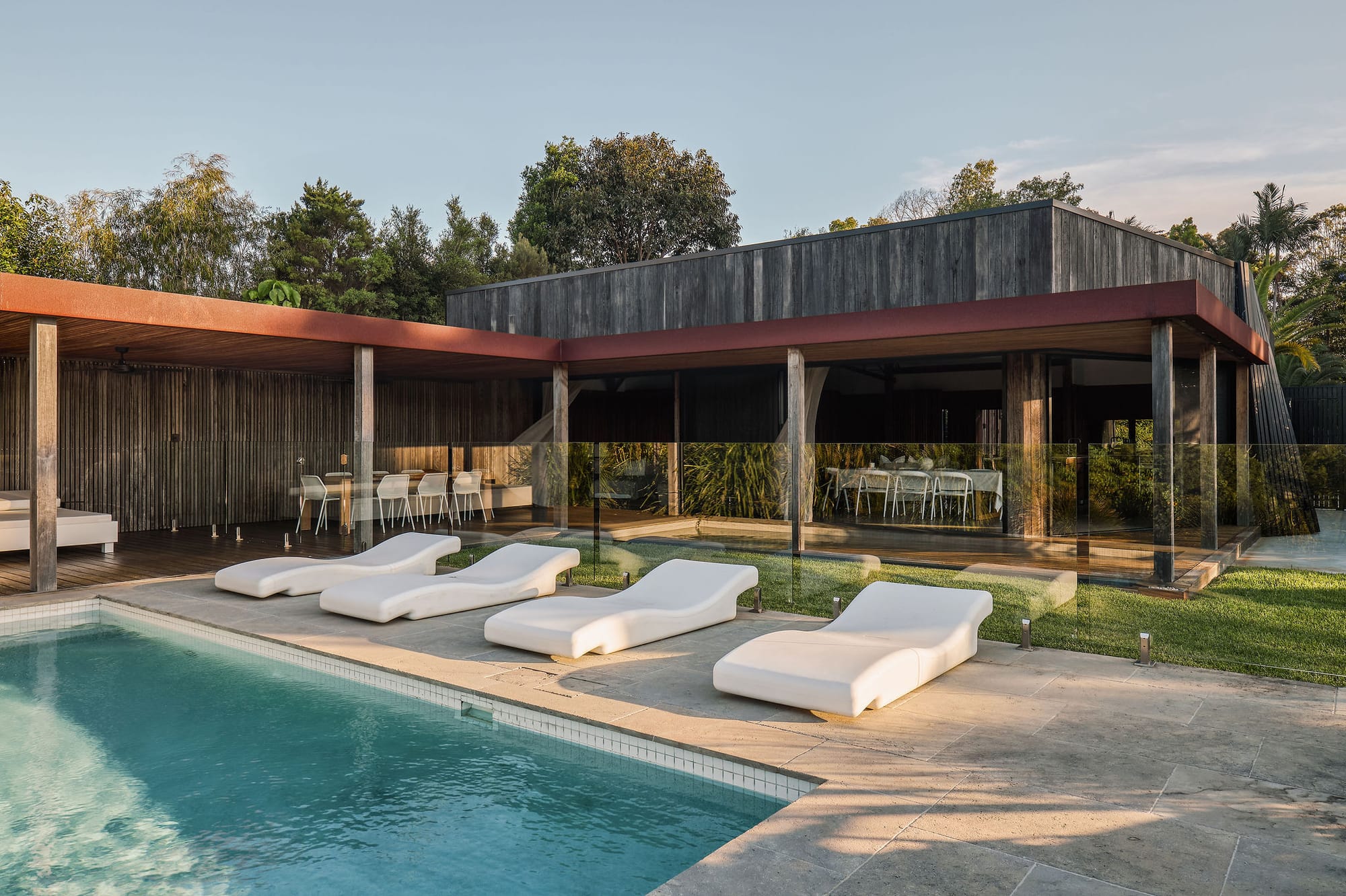 Rockpool Farm Byron Bay. Photography by Andy Macpherson. White recliner lounges by pool. Timber deck wraps around home behind glass pool fence.