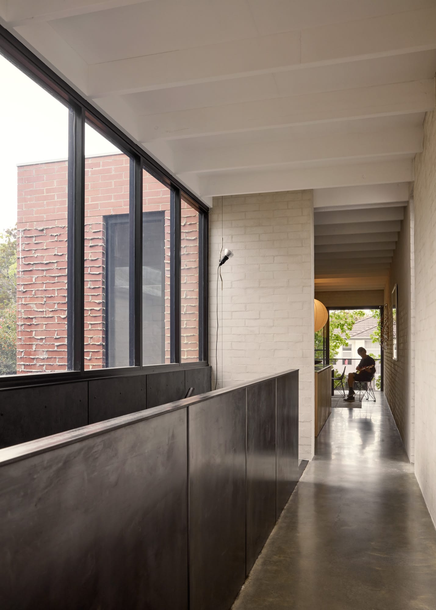 Brick House by Studio Roam. Photography by Jack Lovel. Metal stair balustrade, polished concrete floors through hallway. White brick walls. Windows overlooking street at end of hall. 