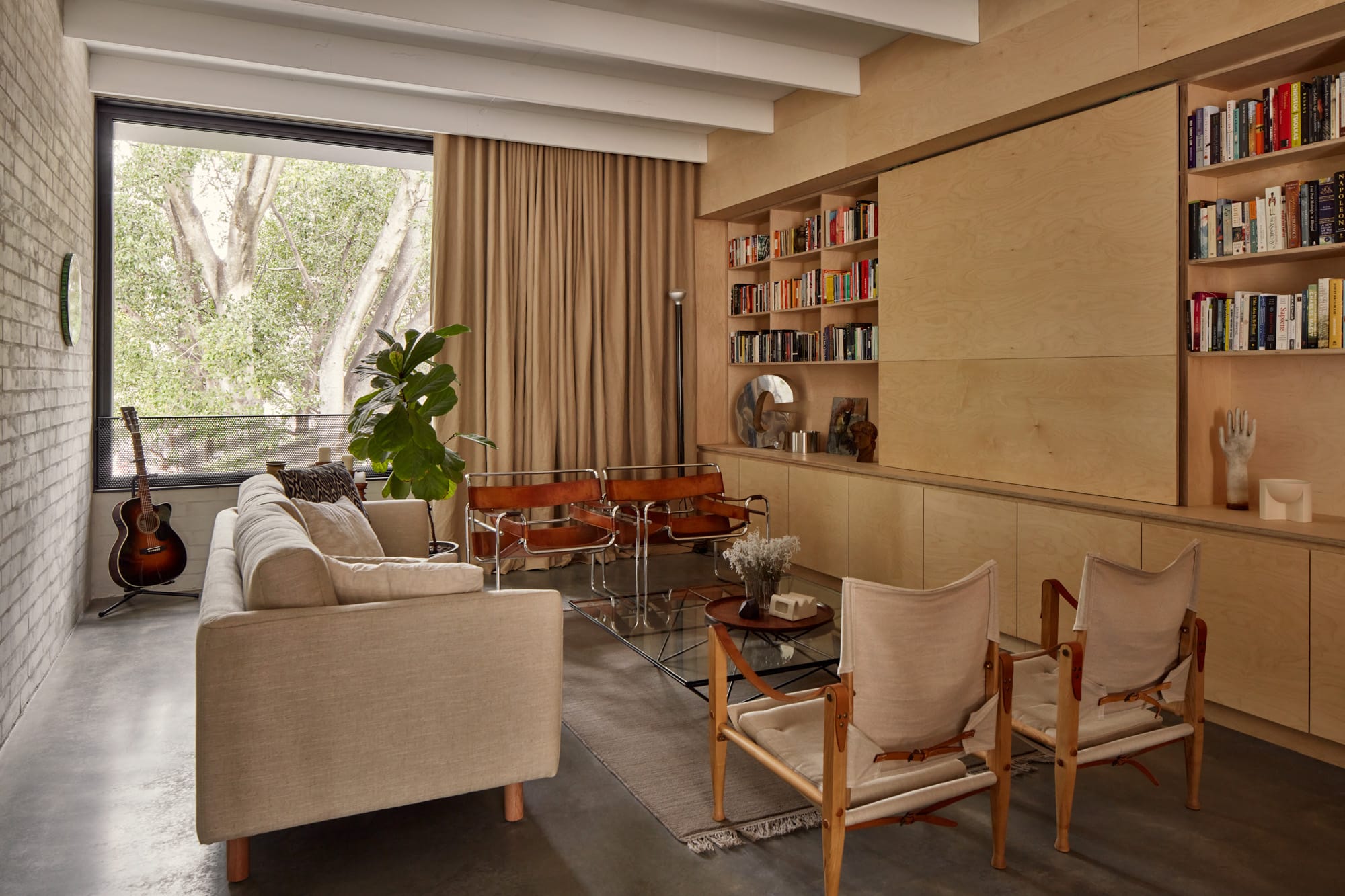 Brick House by Studio Roam. Photography by Jack Lovel. Living space with floor to ceiling plywood storage and bookshelf on right hand side. Eclectic armchairs and couch. Polished concrete floors. 