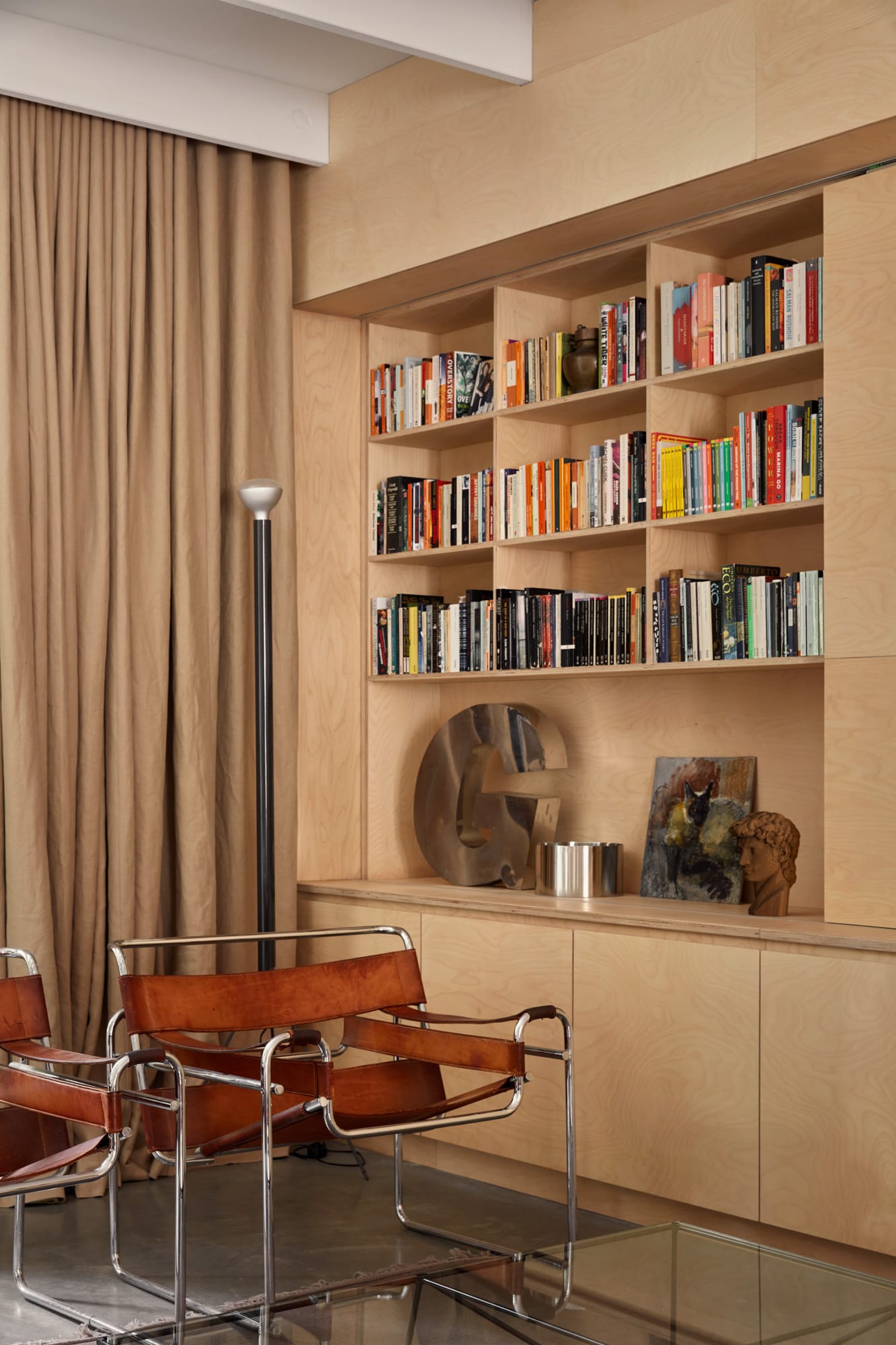 Brick House by Studio Roam. Photography by Jack Lovel. Floor to ceiling plywood bookcase and storage to left of image. Chrome and tan leather armchairs on polished concrete floor. 