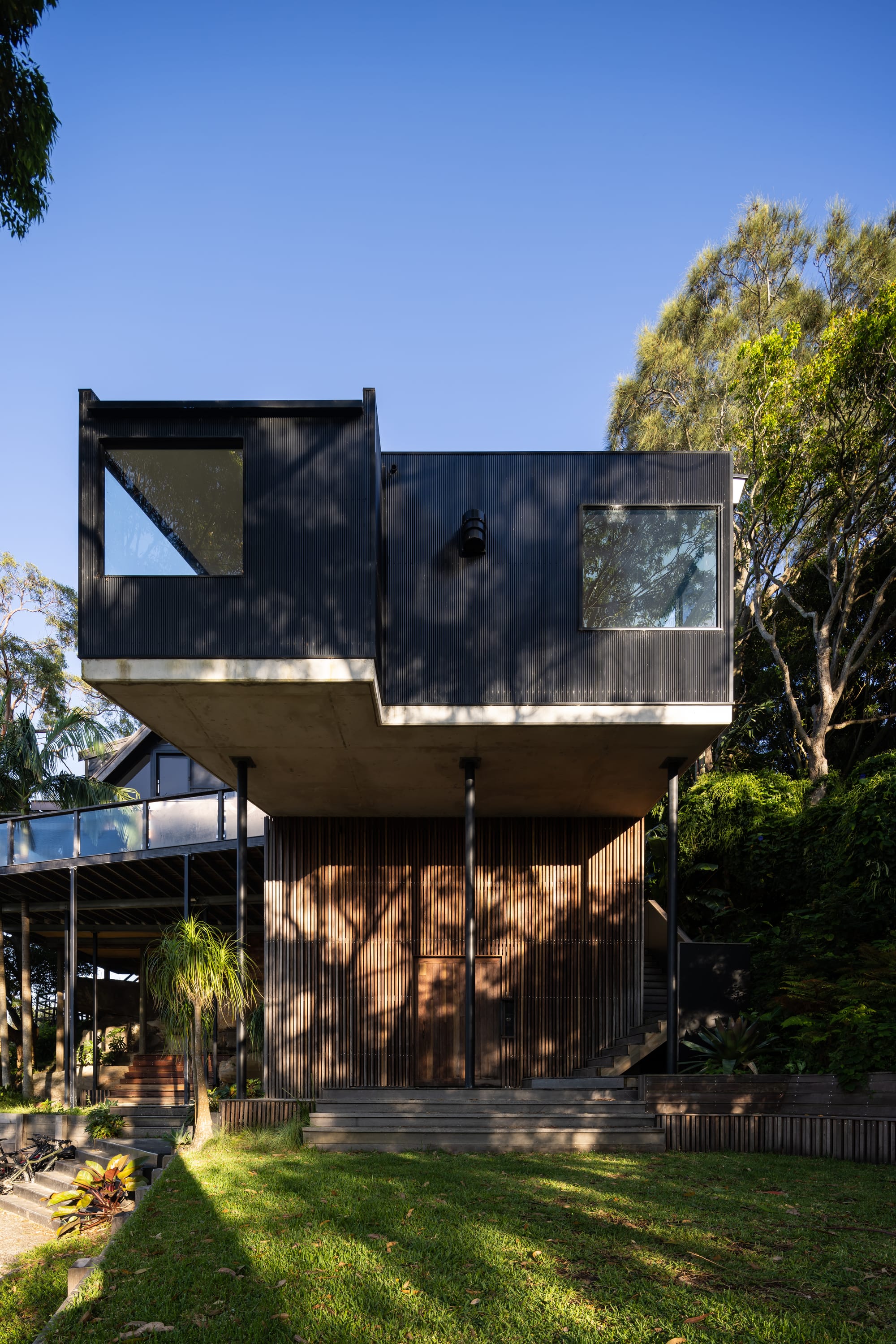 Tree House by North By North. Photography by Simon Whitbread. Facade of double storey home with raw timber clad on ground floor and black clad on second storey. Green front lawn. 