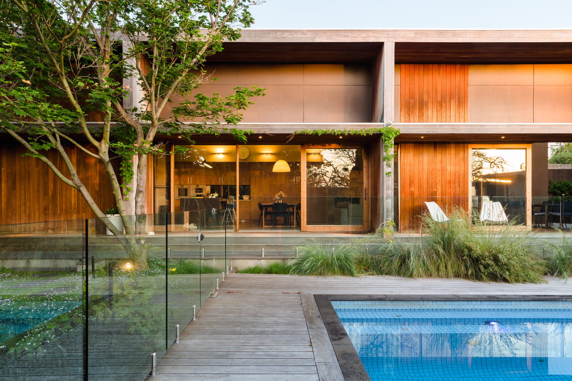 Wills Kew by Julie Crowe Landscape Design. Photography by Erik Holt Photography.  Long double storey home with pool at the front. Timber decking and glass fencing around pool. Plants growing through decking and along second floor of home. 