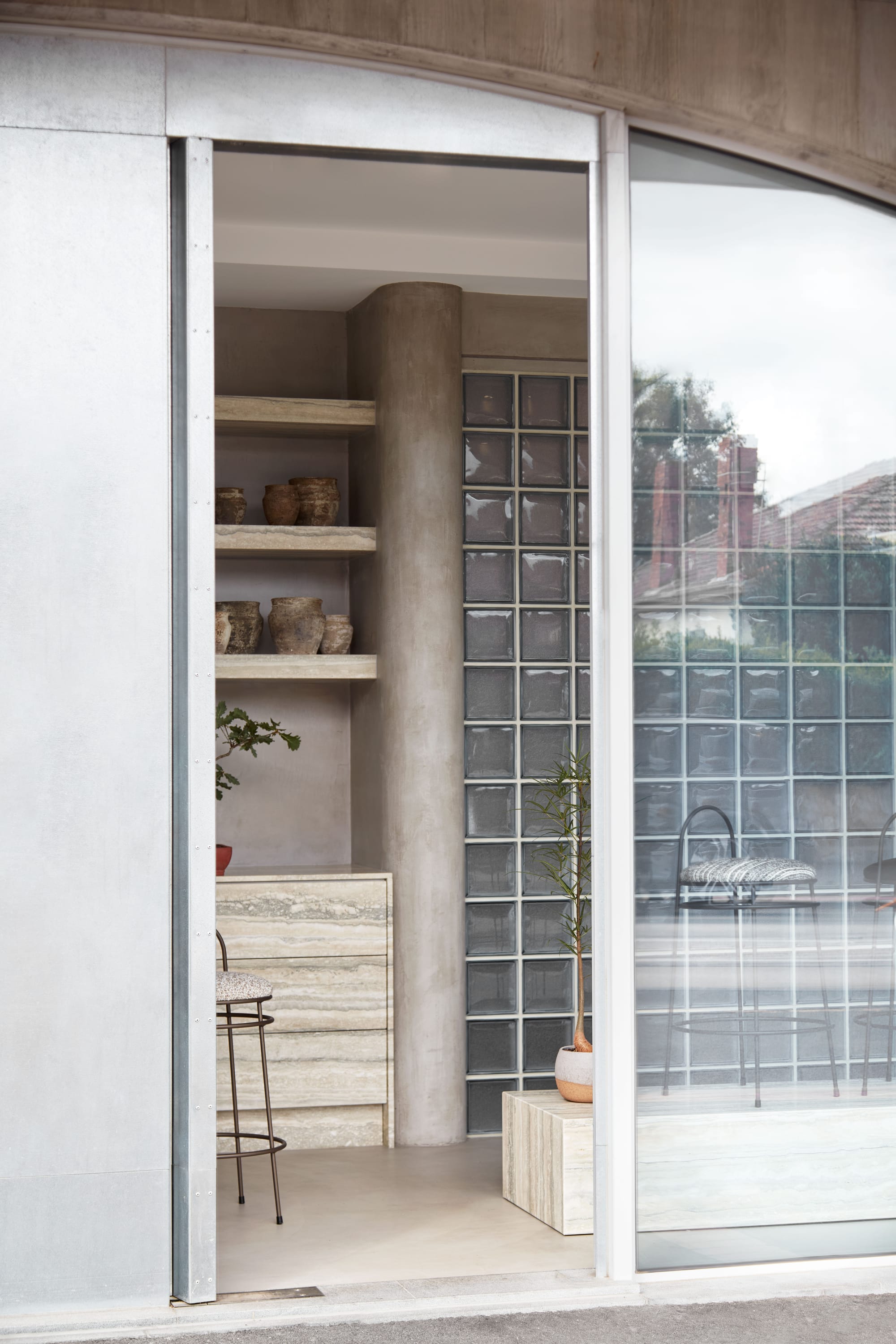 Zenn Showroom. Photography by Elisa Watson. Doorway from outside leading inside. Glass brick wall and shelving visible through doorway. Small potted plants on shelves and podiums. 
