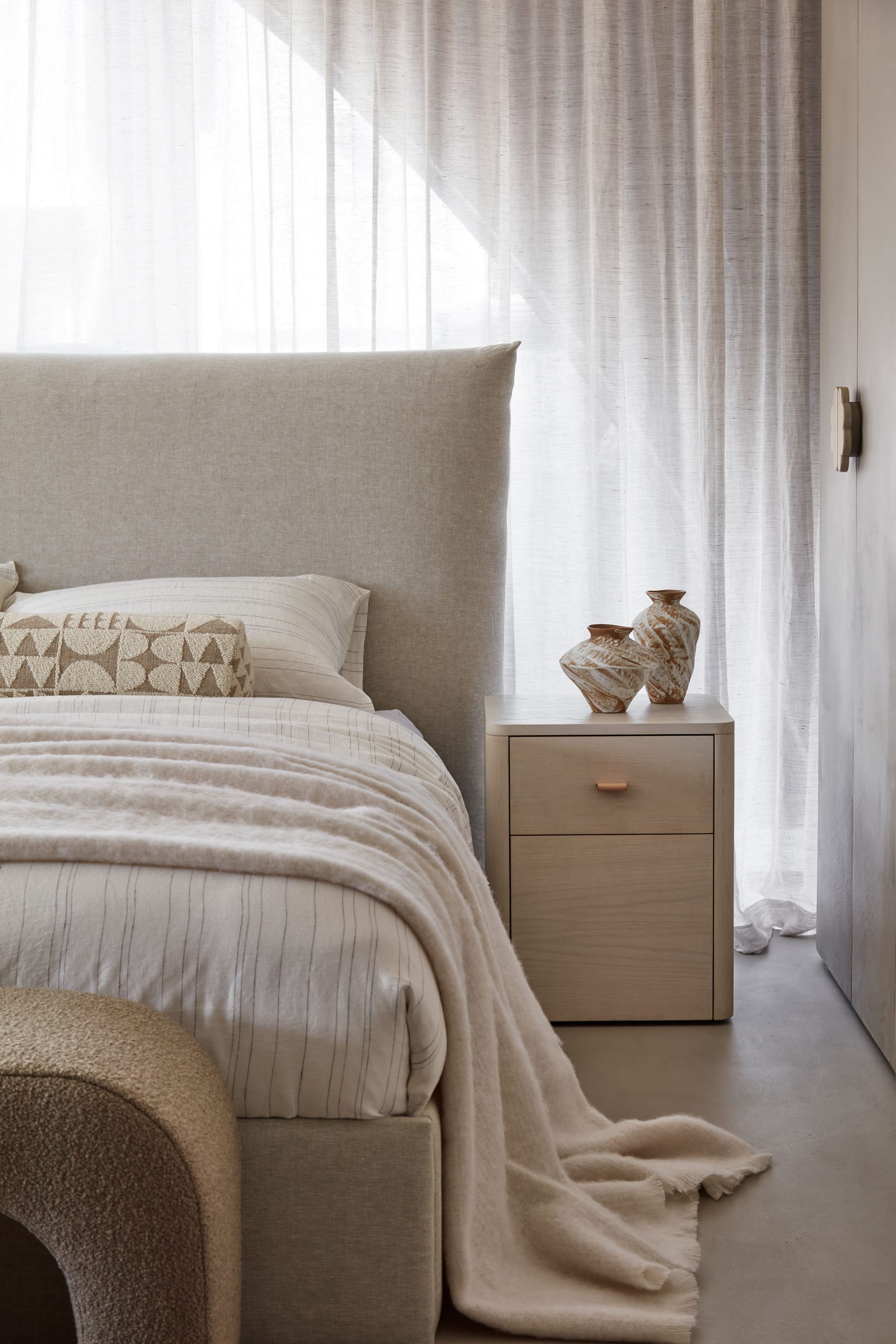 Zenn Showroom. Photography by Elisa Watson. Bed with upholstered headboard and neutral linen bedding. Light timber bedside table. Floor to ceiling white sheer linen curtains.  