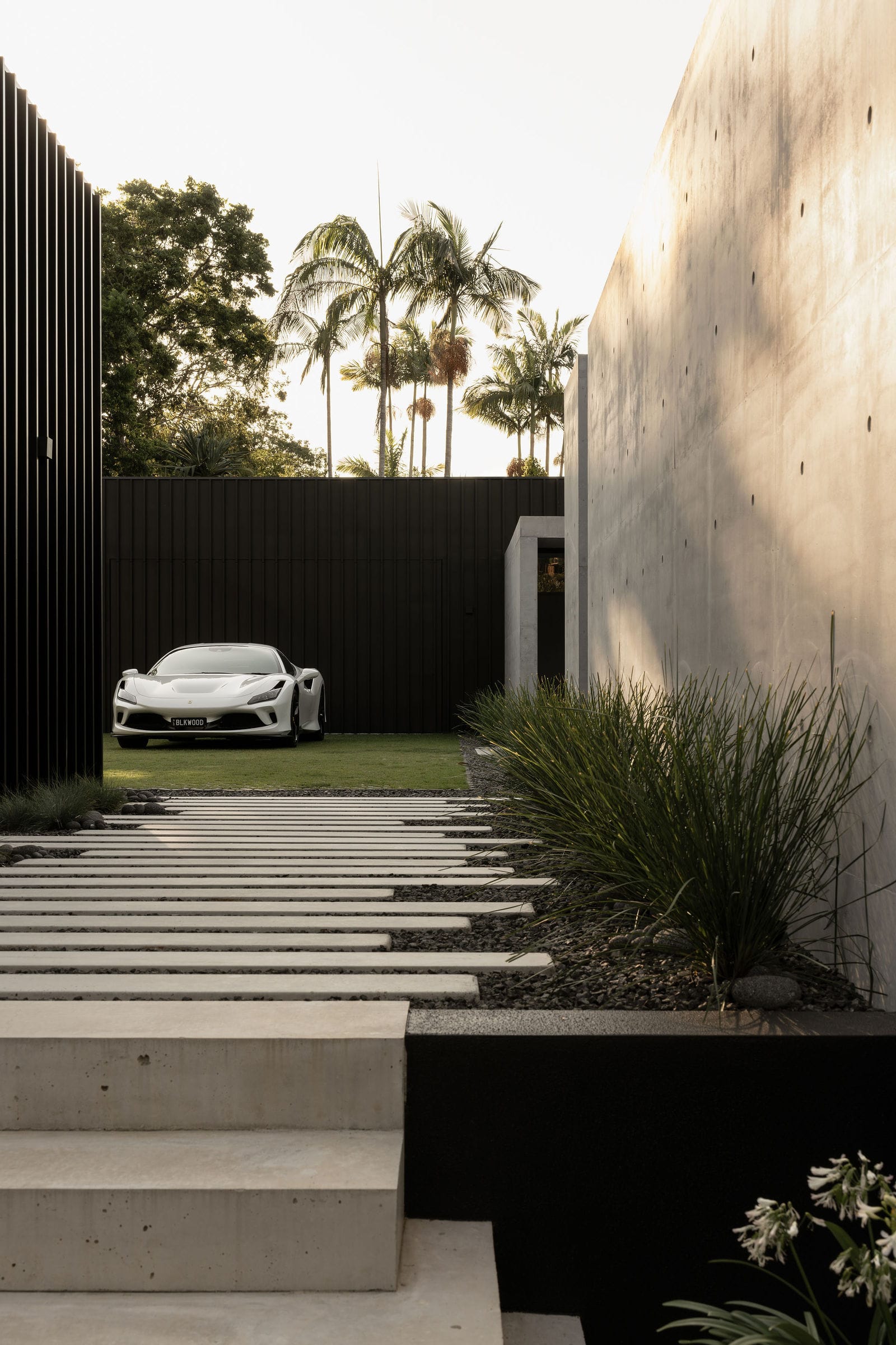 Blackwood by Sarah Waller Architecture. Photography by Alyne Media. Outdoor steps into enclosed grass space with white sportscar. Concrete walls. 