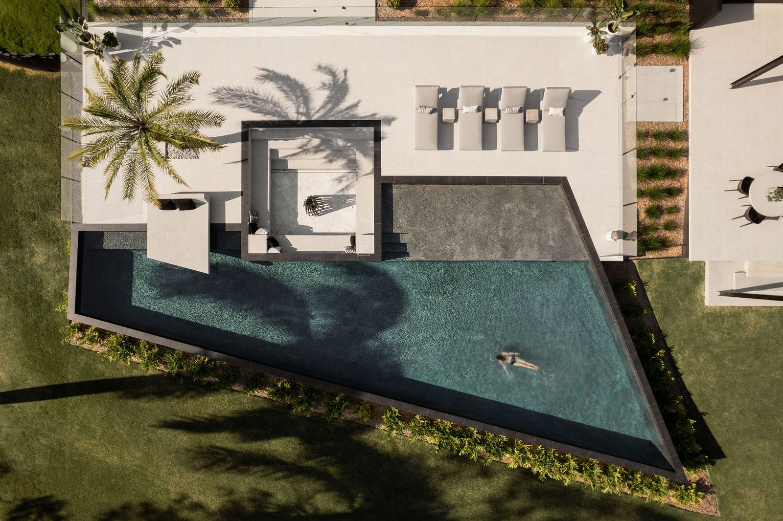 Blackwood by Sarah Waller Architecture. Photography by Alyne Media. Birds eye view of angled pool with concrete patio with firepit, concrete day bed and chaise lounges. 