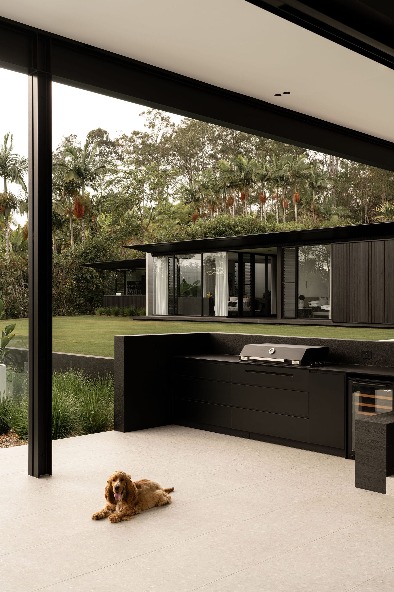 Blackwood by Sarah Waller Architecture. Photography by Alyne Media. Outdoor kitchen with concrete floor tiles and black kitchen cabinetry and barbeque. Blac timber clad pavilion home in background with large grass area at front. 